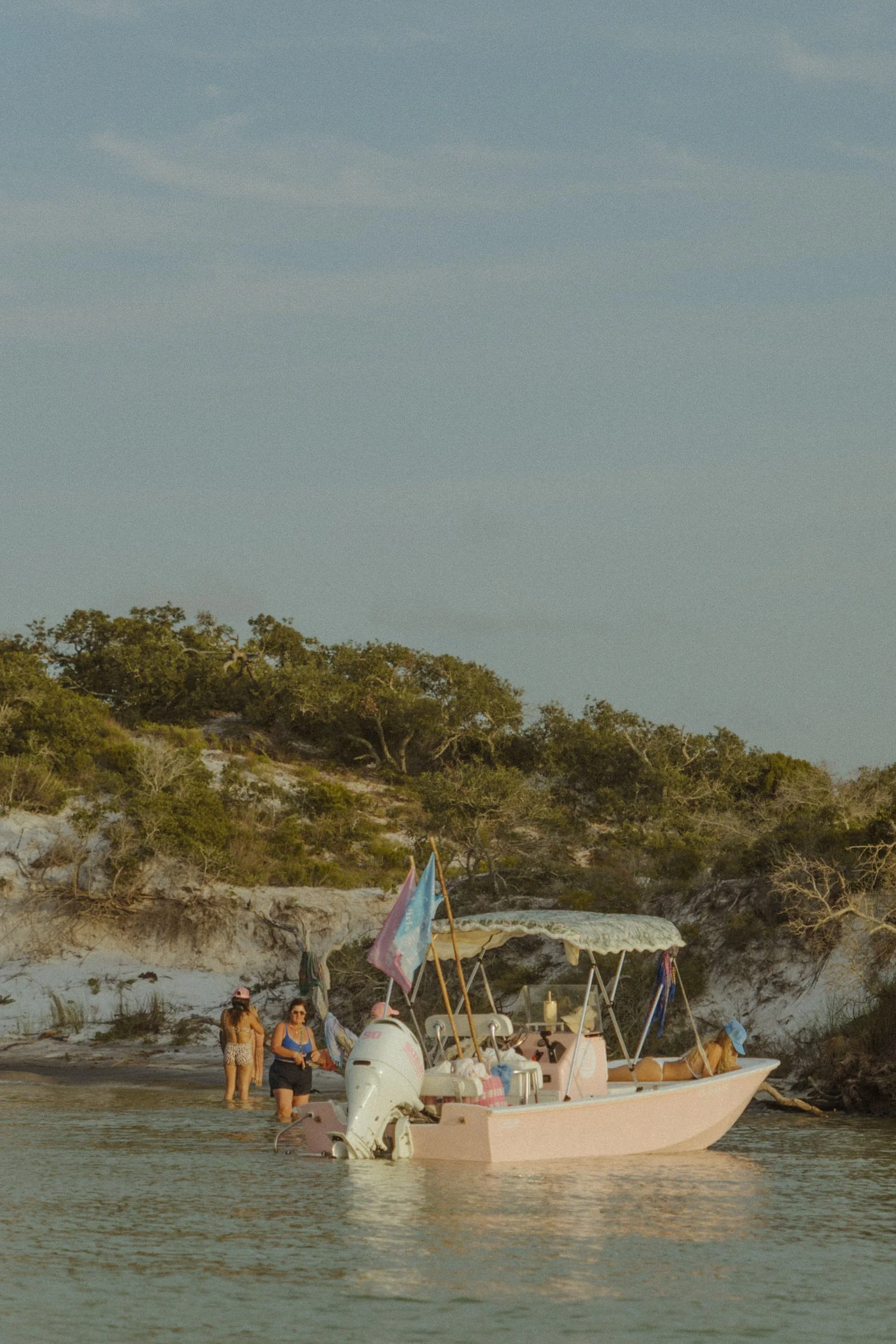 Three women standing in shallow water near a pink boat with a canopy at a beach with a rocky shoreline and trees in the background.