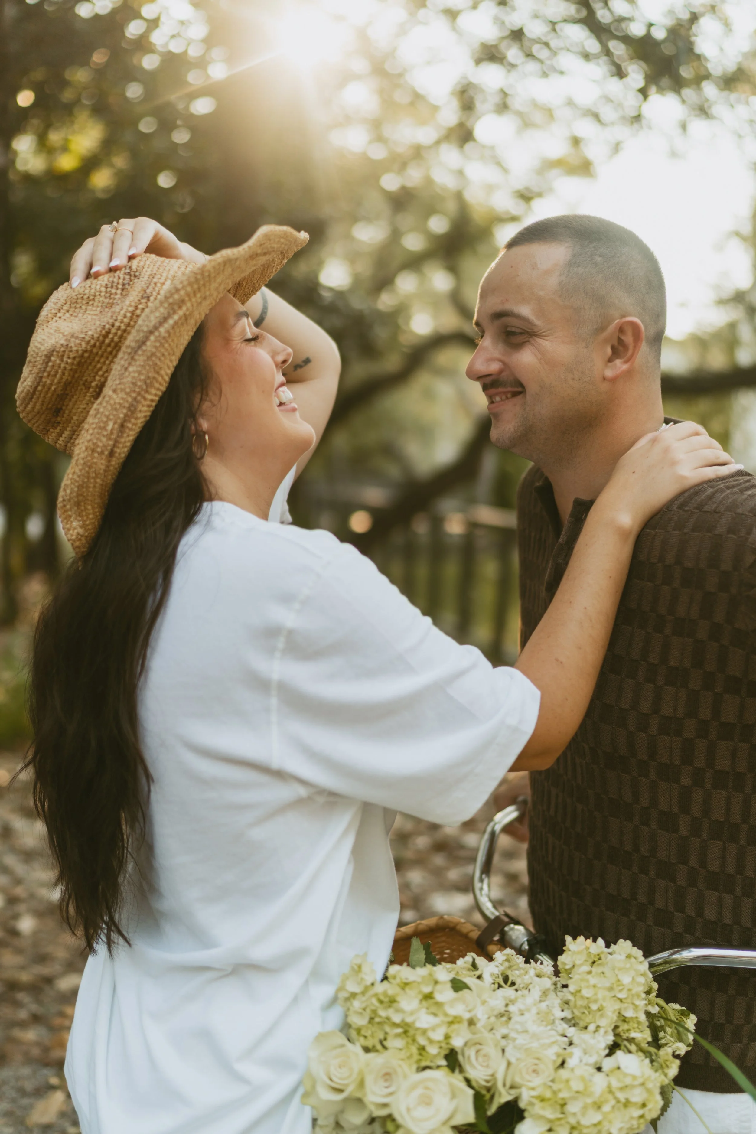  couple laughing together for engagement photos, he is looking at her smiling and she is laughing with a hand on his shoulder and one on her hat leaning over the bike  