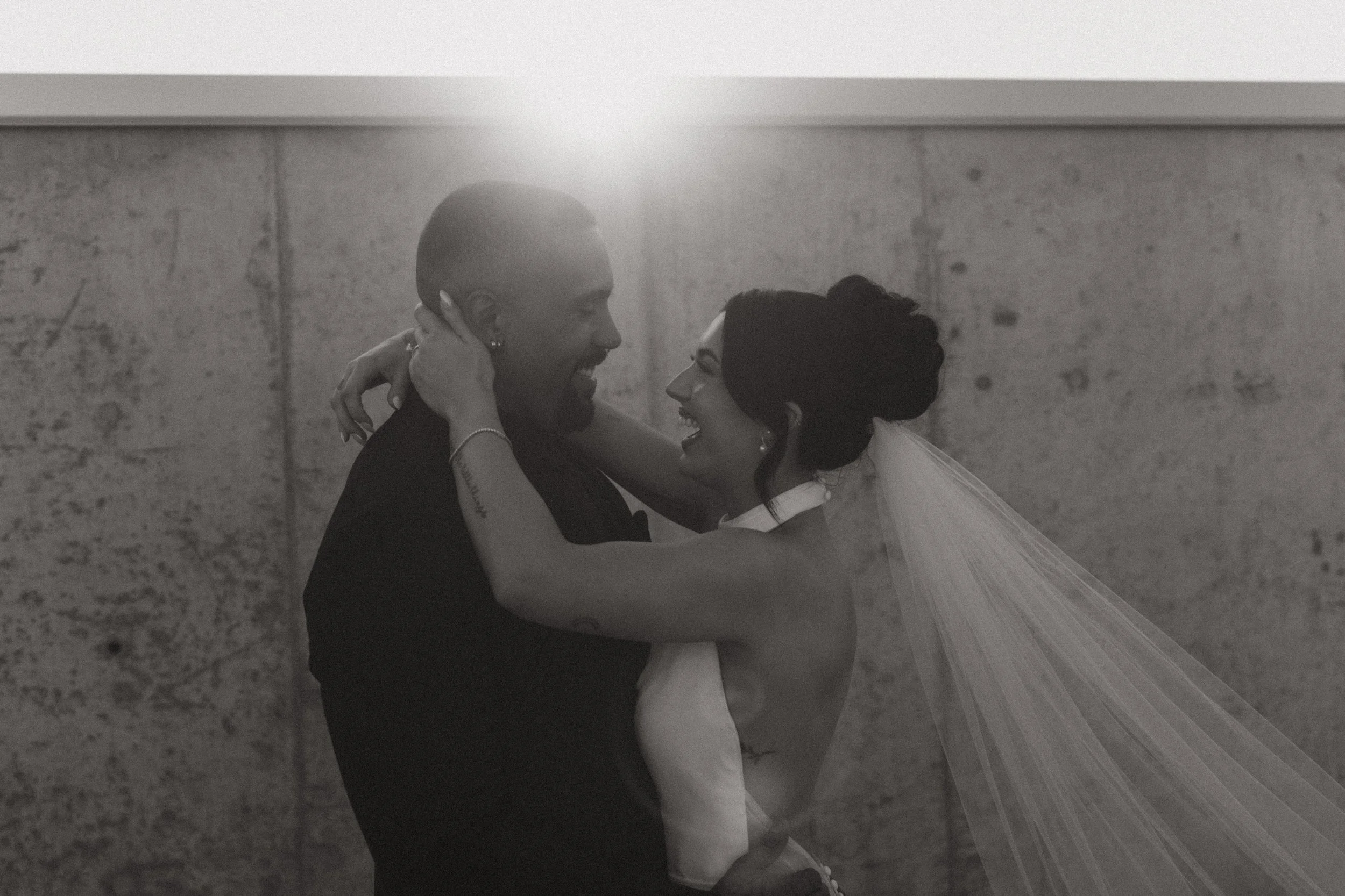bride and groom laughing together in a black and white photo while her veil blows in the wind and the sun shines from behind them on the roof of a parking garage
