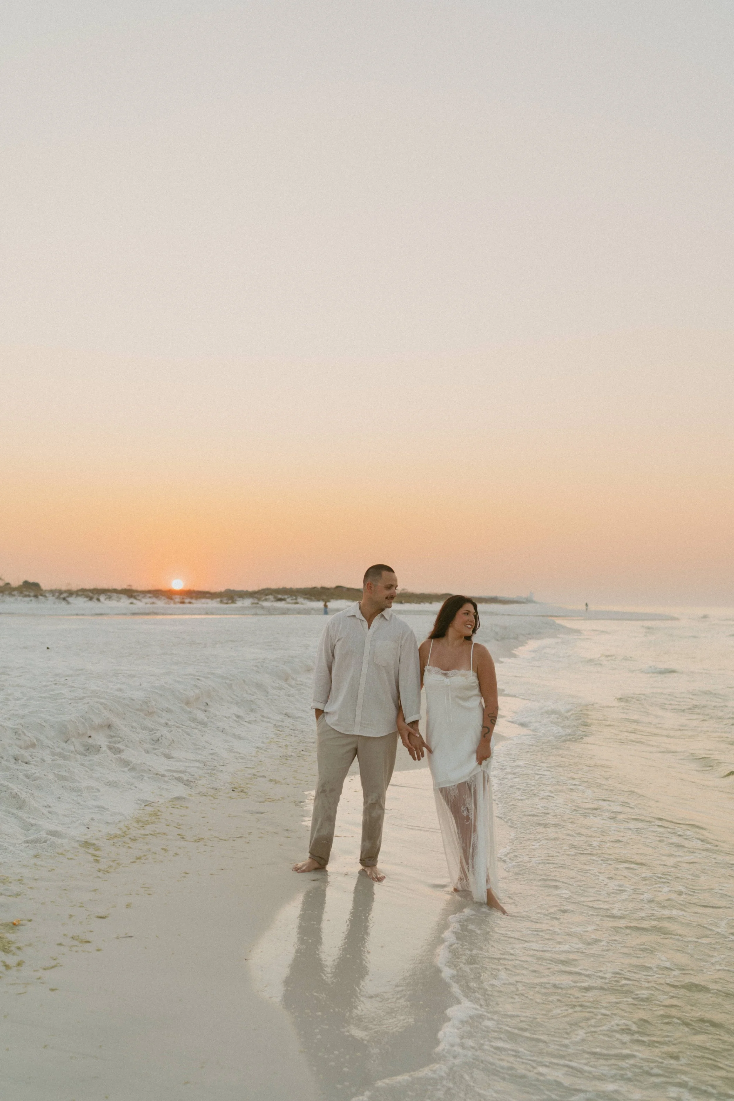  couple looking out to the water at sunrisse for engagement photos, they are holding hands, she is looking at the water and he is looking at her the sun is rising behind them over the dunes, the sky is glowing orange and yellows the water is calm and