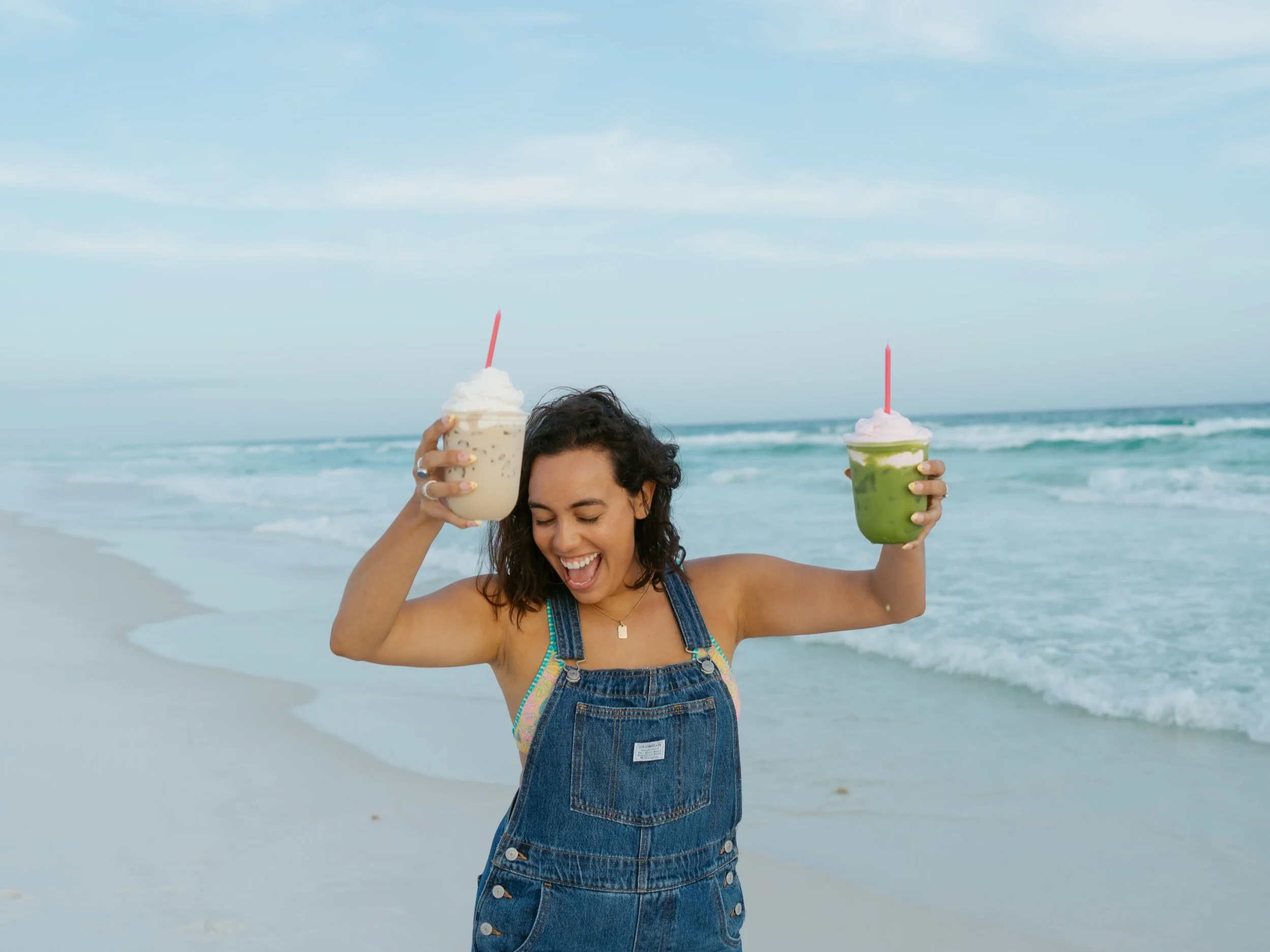 girl in overalls and a bikini holding a latte and a matcha in each hand dancing around excited celebrating on the beach