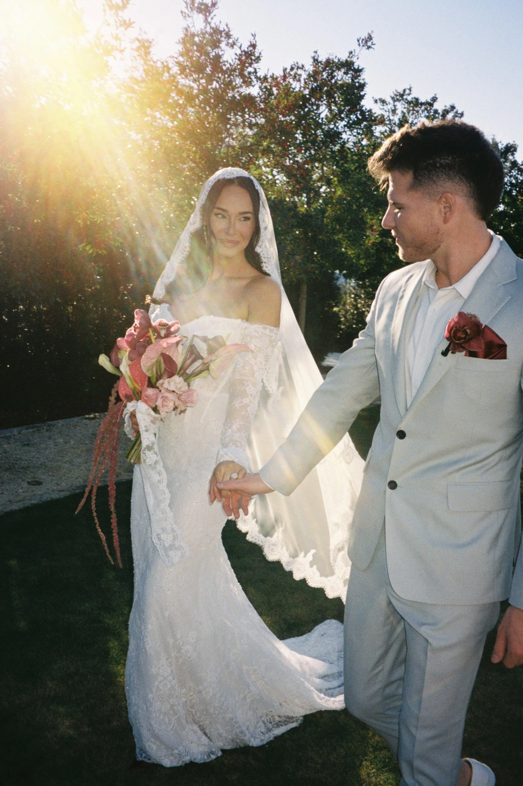 A bride and groom hold hands outdoors during sunset, with the bride in a white lace wedding dress and veil holding a bouquet, and the groom in a gray suit with a pink boutonniere.