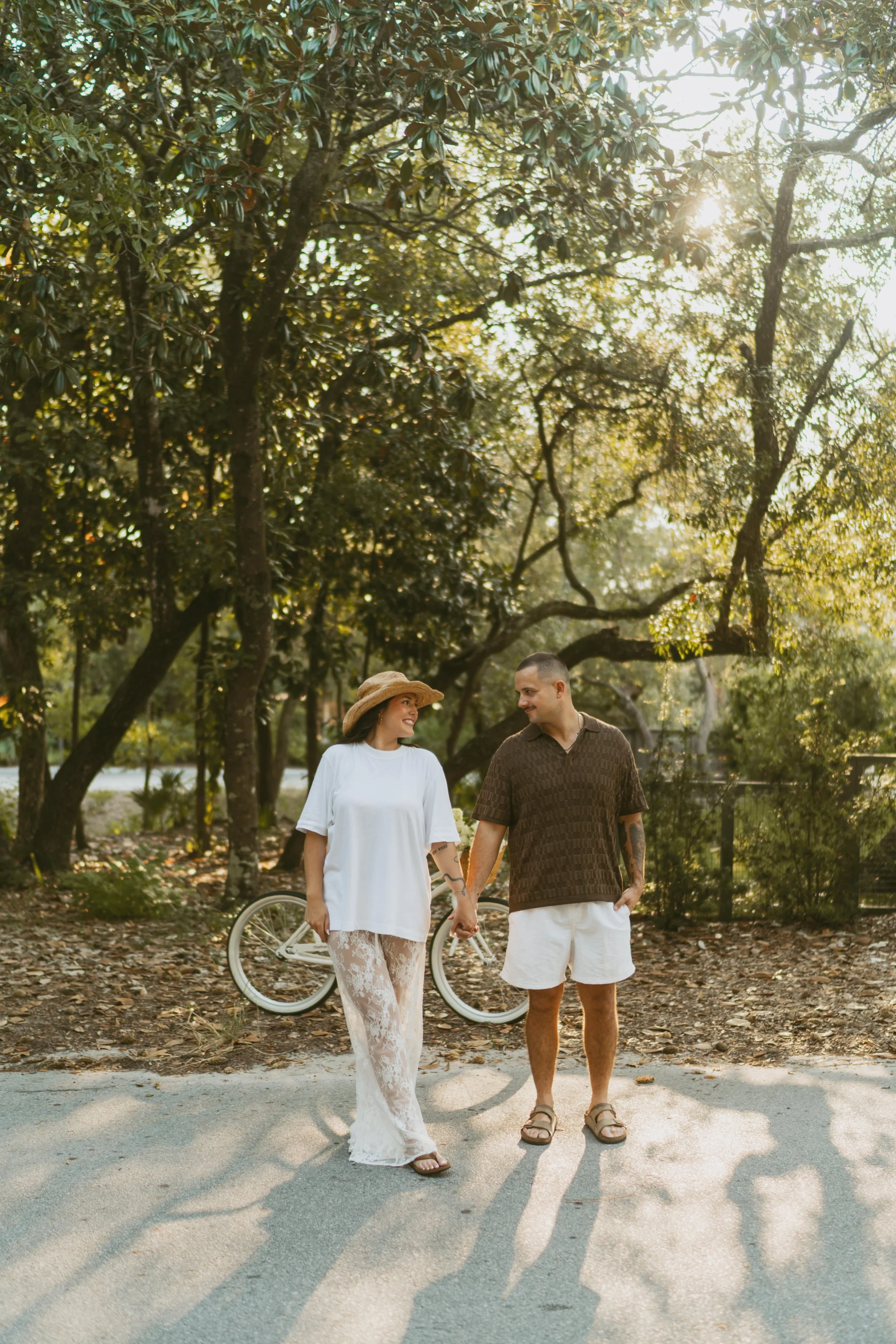  couple looking at each other holding hands on the side of the street with the trees lined up behind them  
