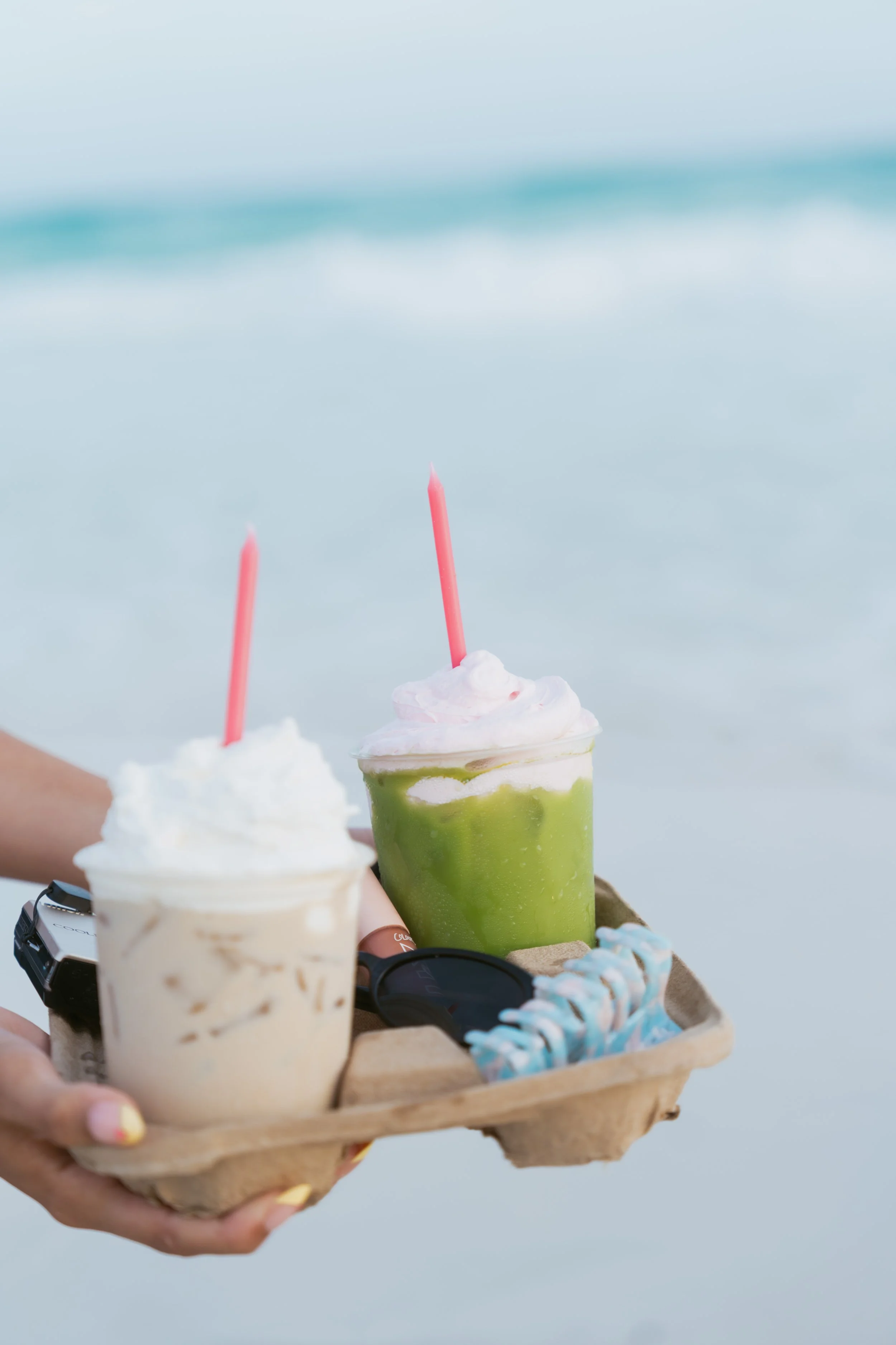  latte and matcha in a drink carrier on the beach with a hair clip, sunglasses, lip gloss, and a digital camera  