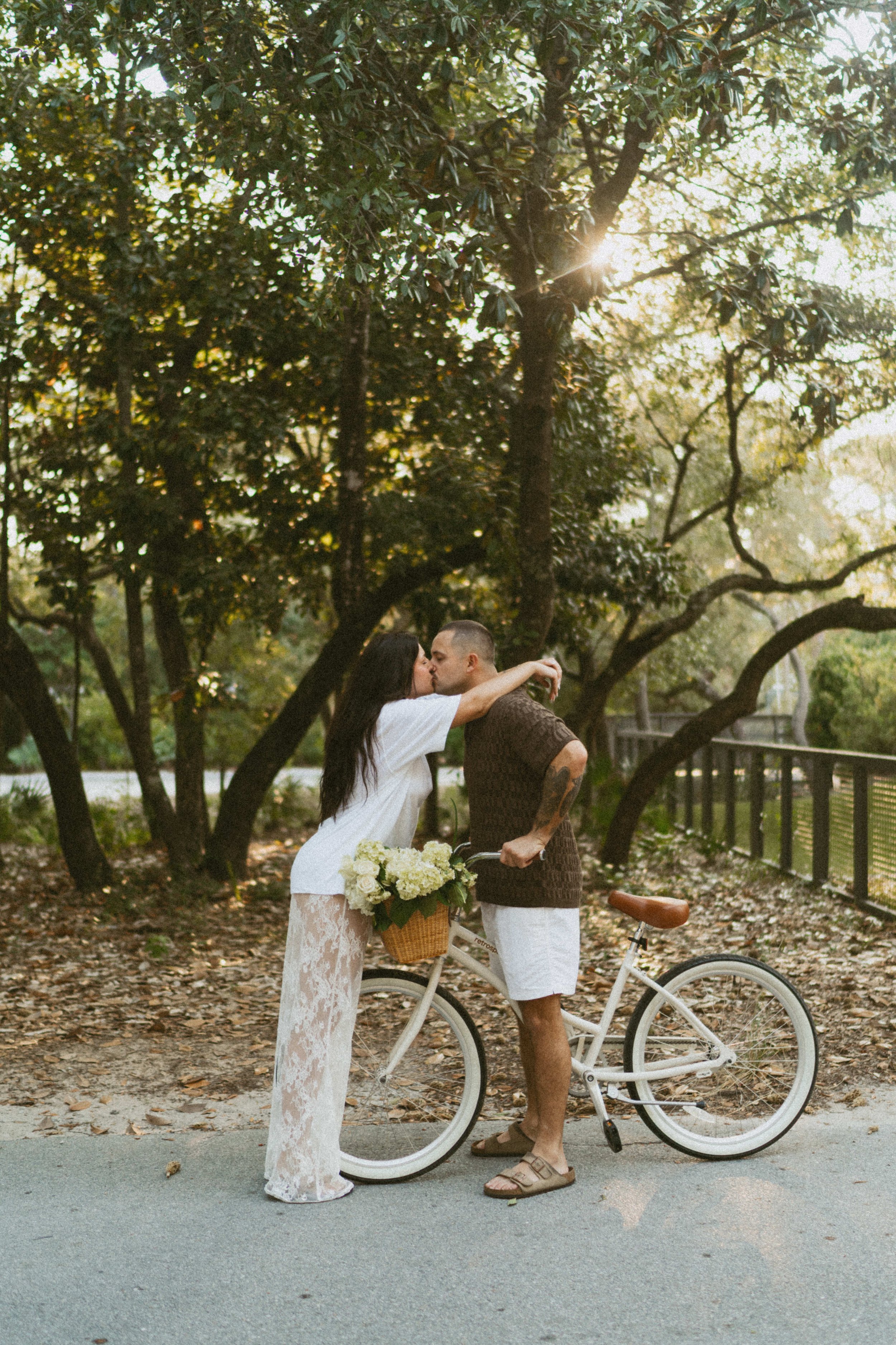  couple kissing on bikes for engagement photos, he is on a white bike with a basket and she is standing over the front with her arms over his shoulders kissing him the bike has a basket with flowers in it and the sun is poking through behind the tree