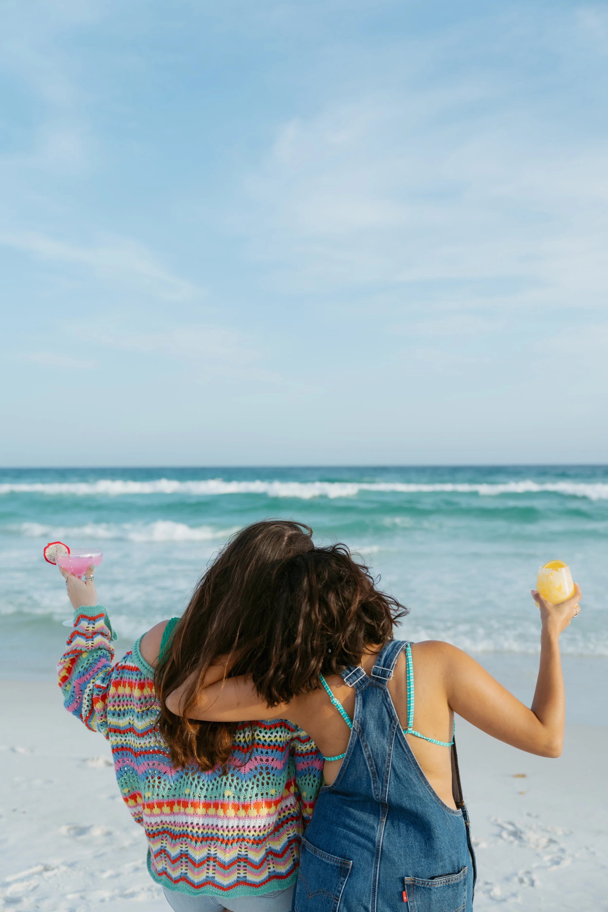  two girls leaning on each other looking out at the gulf of mexico holding their drinks up  