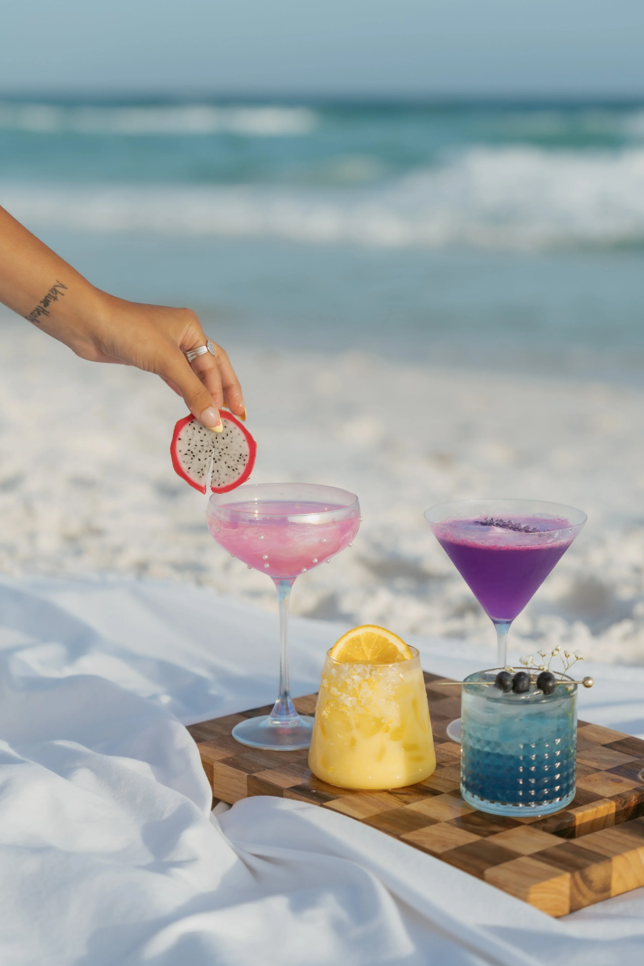  4 drinks on a cutting board on the beach being with hand adding the toppings  