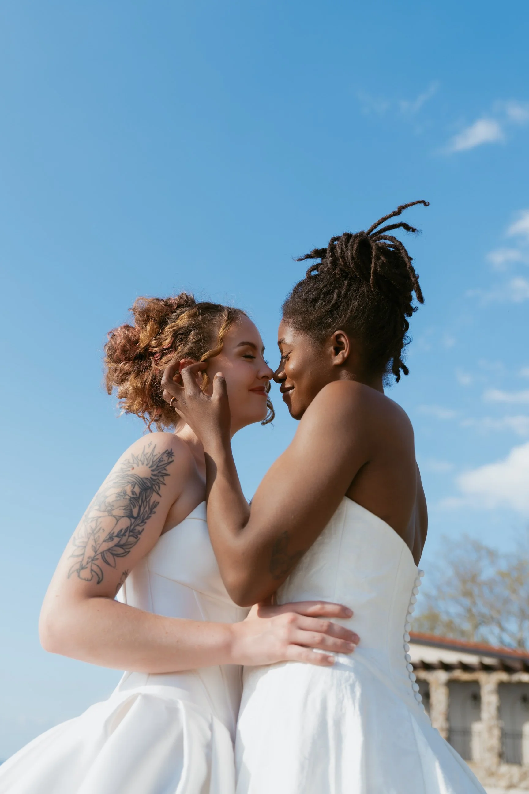 Two women in wedding dresses sharing an intimate moment outdoors with a blue sky background.