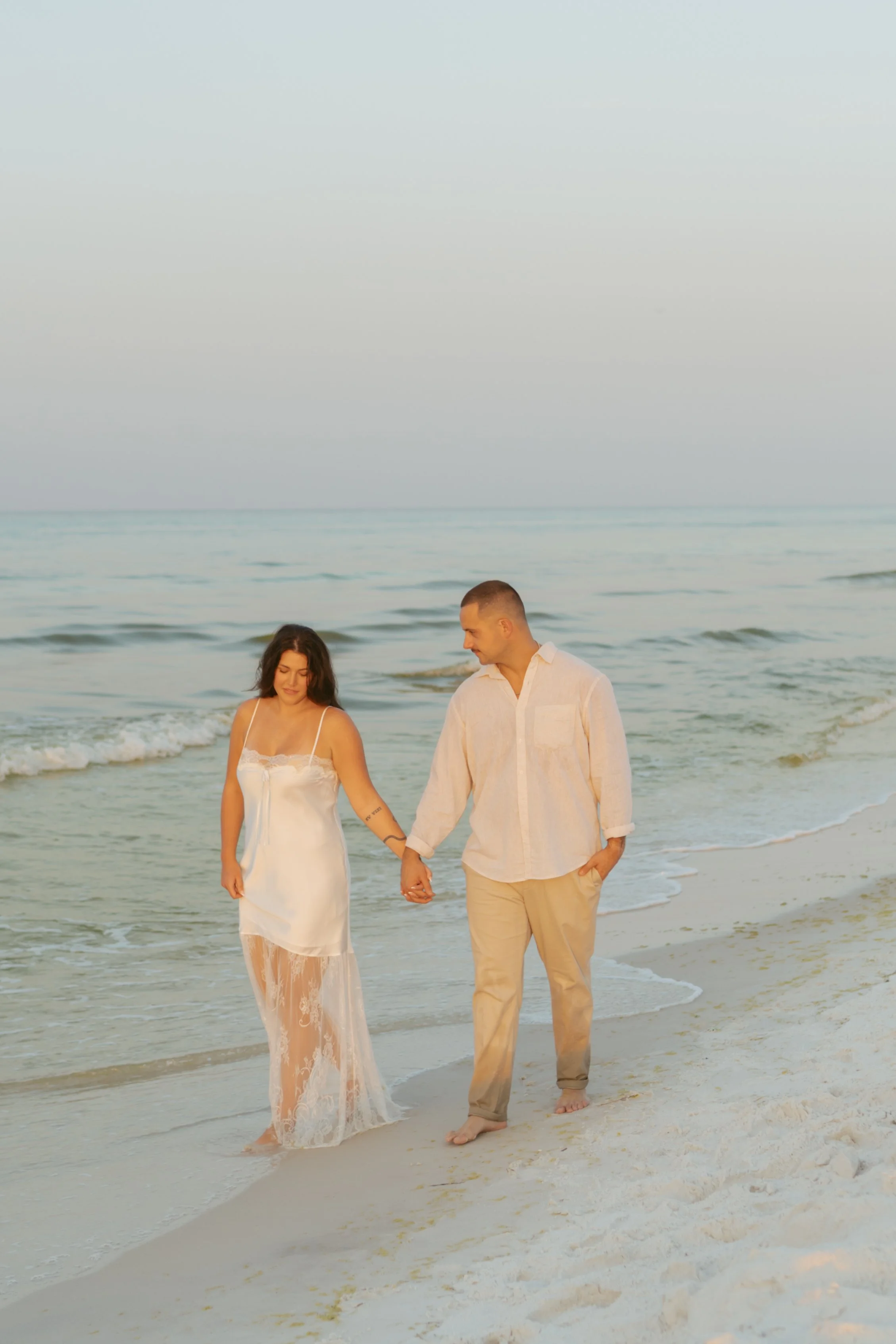  couple walking along the beach at sunrise for their engagement photos she is wearing a white slip dress that has a lace skirt, he is wearing tan pants and a white long sleeve button up they are holding hands she is looking down at the sand and he is