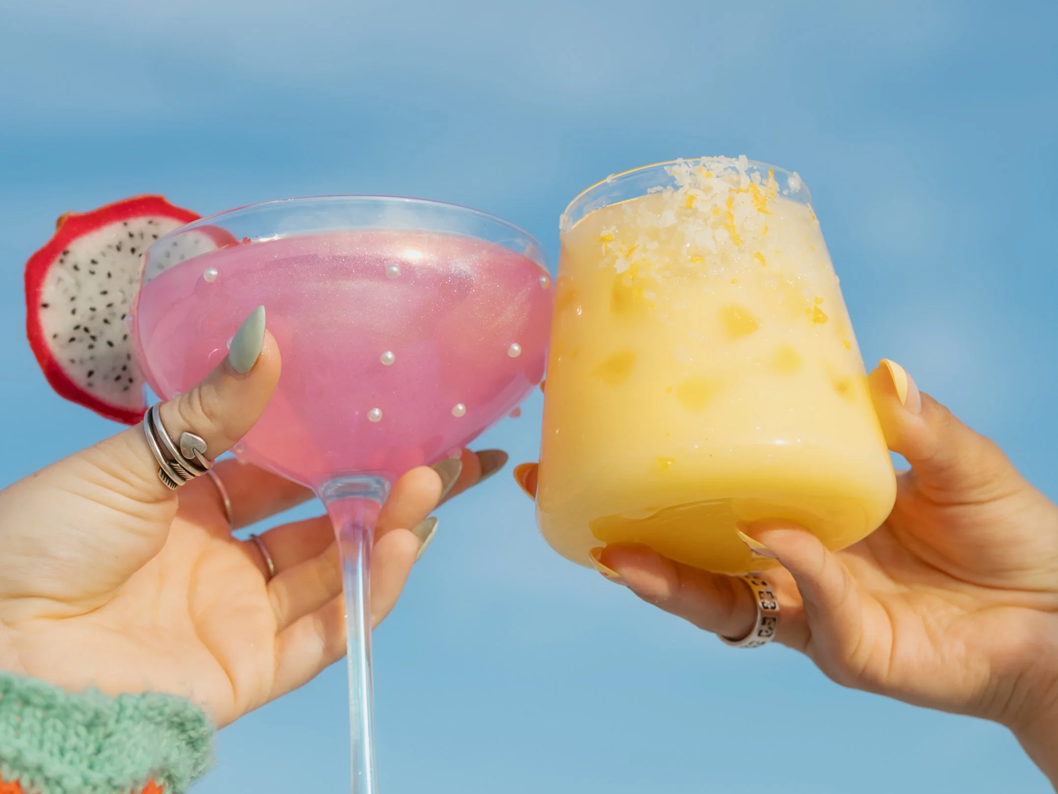hands holding drinks on the left is a pink sparkly drink in a martini glass with pearls and a dragon fruit on the rim, the right is an orange drink with salt and orange zest on the rim, the girls are cheers-ing the glasses