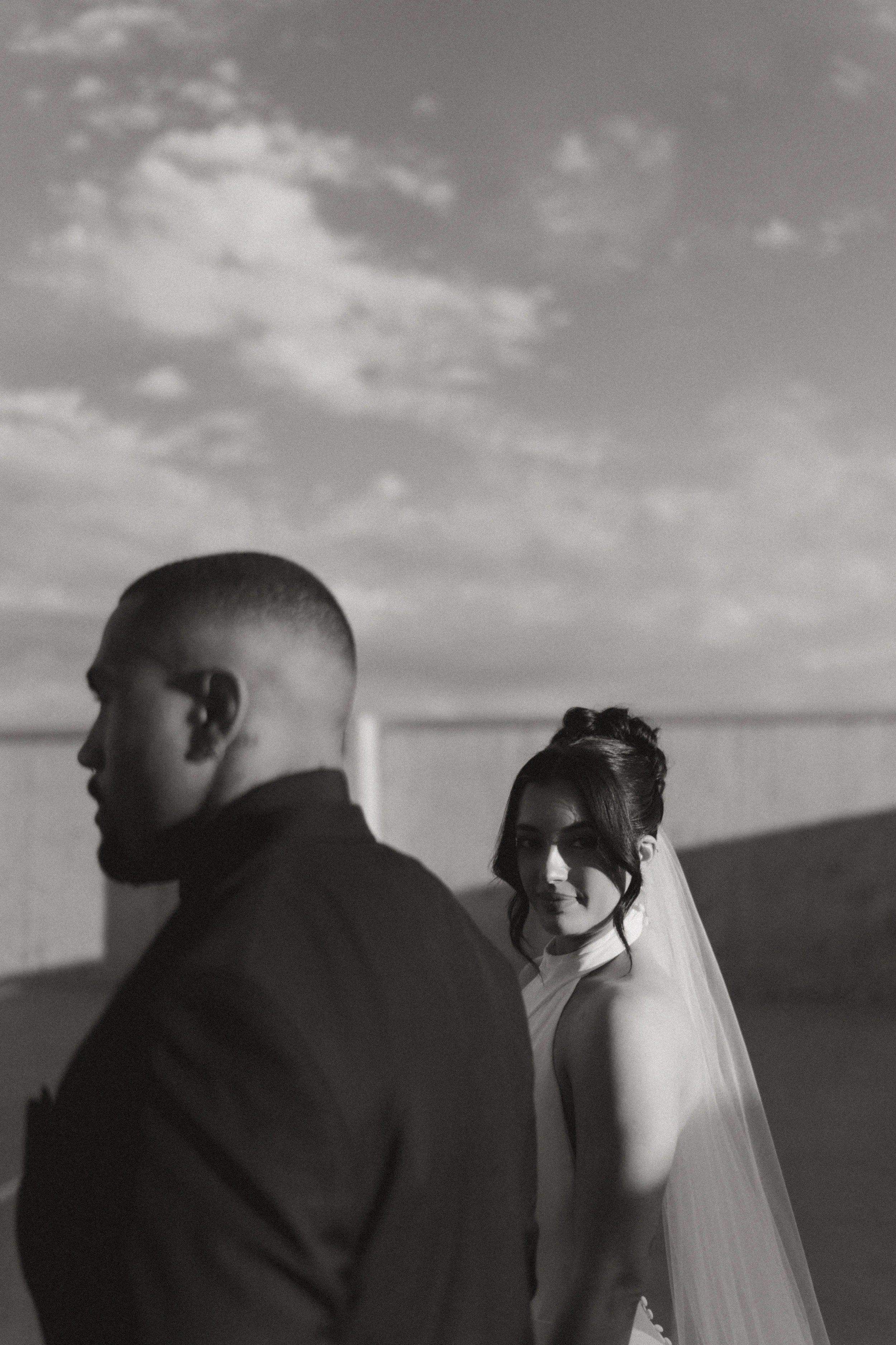  black and white dramatic wedding photo of bride and groom groom looking ahead and bride looking to camera over her sholder while holding her husband’s hand  