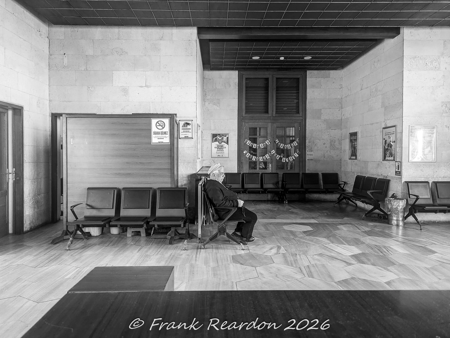 A black-and-white interior view of a passenger waiting room featuring a seated traveler, classic benches, tiled walls, and doors decorated with patriotic bunting.