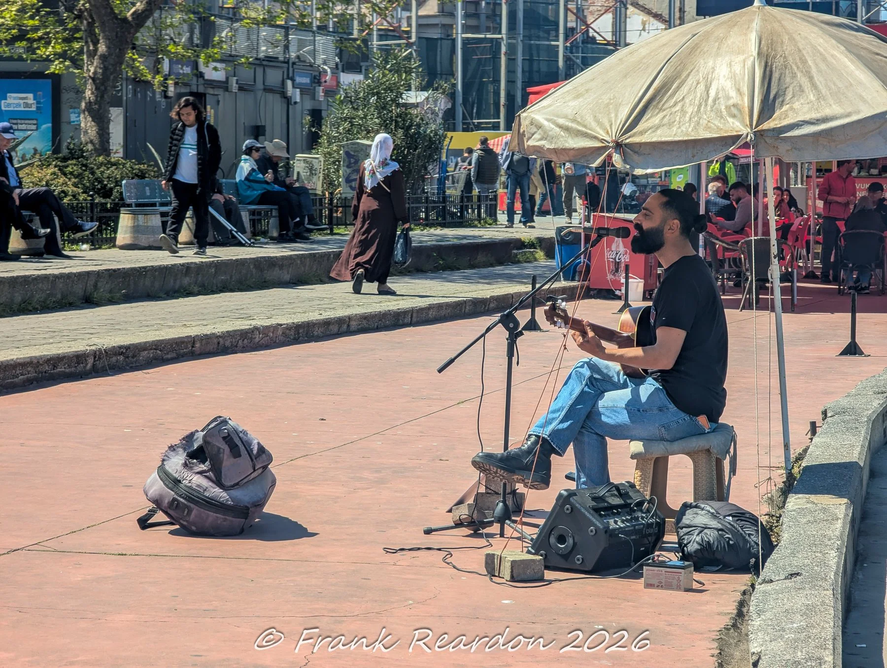 A street performer sitting on a small stool under a beige umbrella, playing an acoustic guitar into a microphone with his amplifier and backpack set up on the pavement.