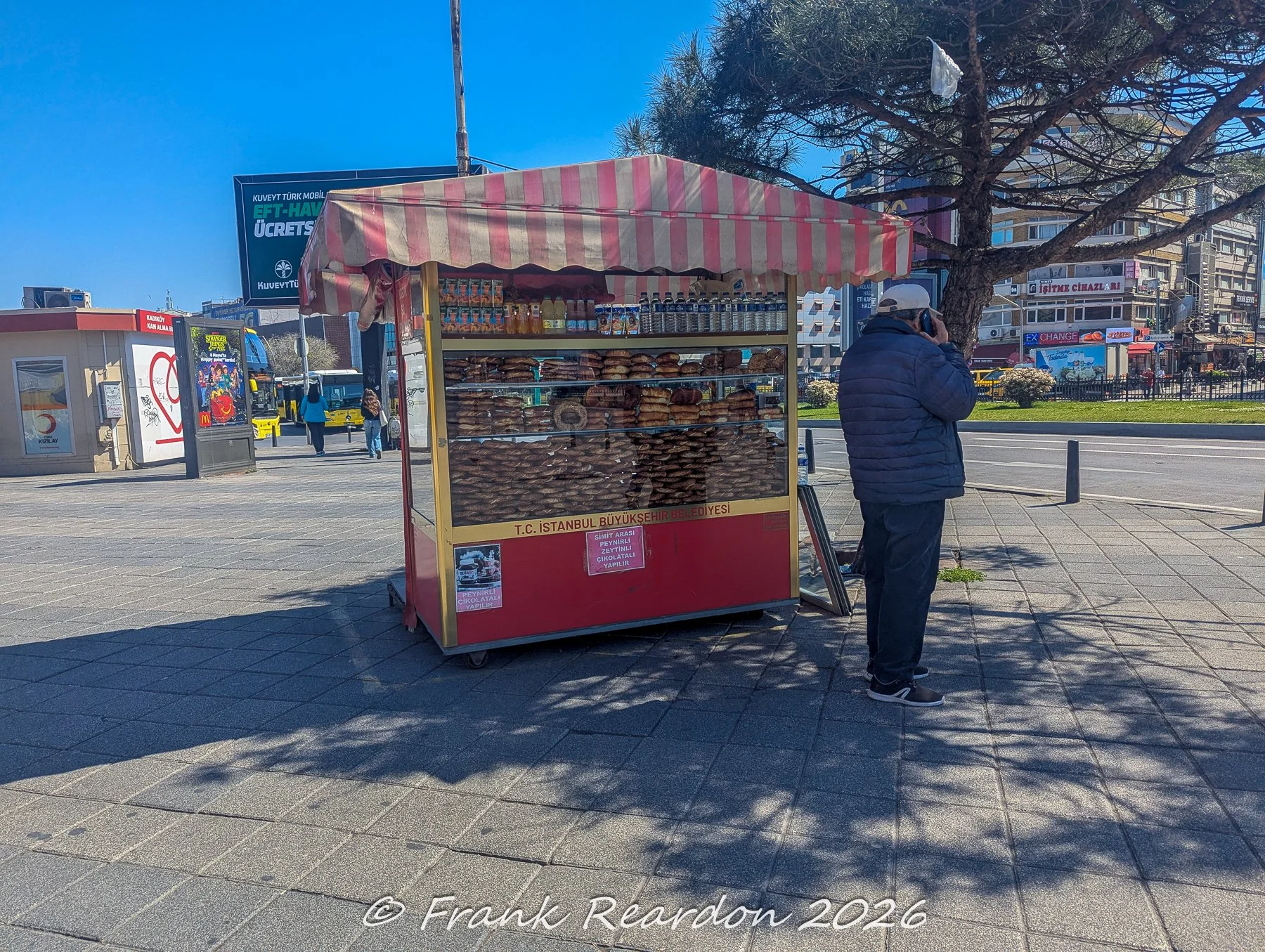 A red-and-white striped street cart operated by T.C. İstanbul Büyükşehir Belediyesi, stocked with stacks of fresh simit bread as a vendor talks on his phone nearby.