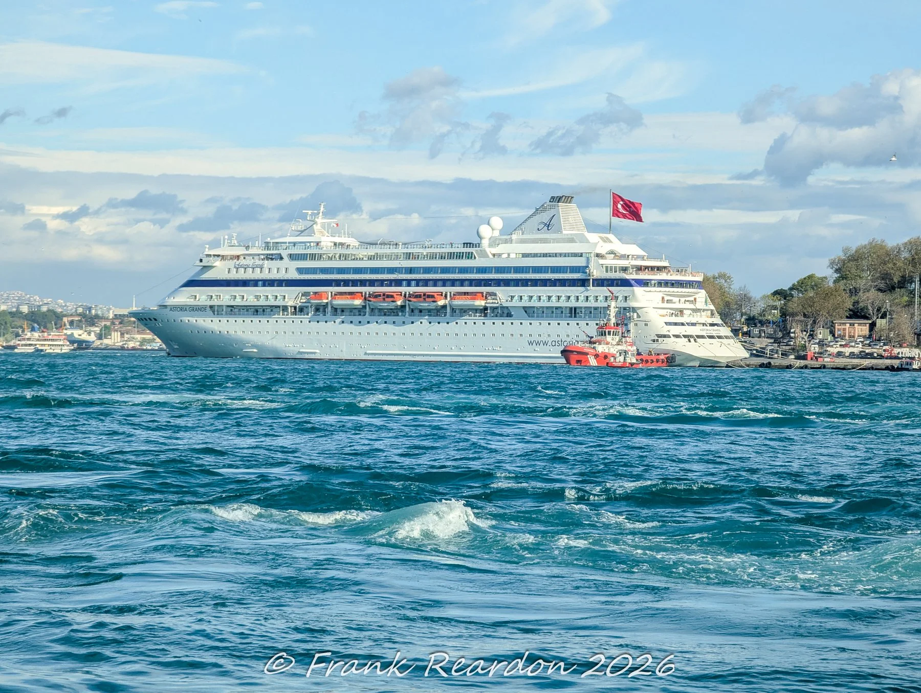 A large white cruise ship, the Astoria Grande, cruising through the waters of the Bosphorus accompanied by a smaller harbor tugboat.