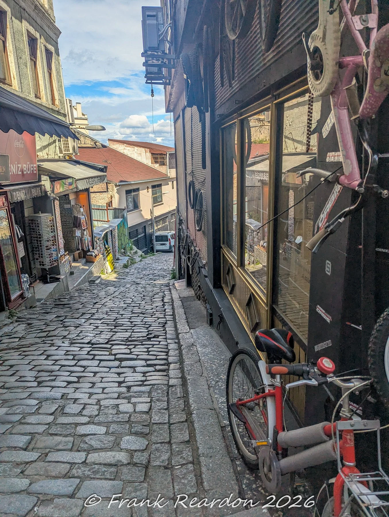 A narrow cobblestone incline flanked by local storefronts, featuring a bicycle shop with a pink bike mounted on the wall.