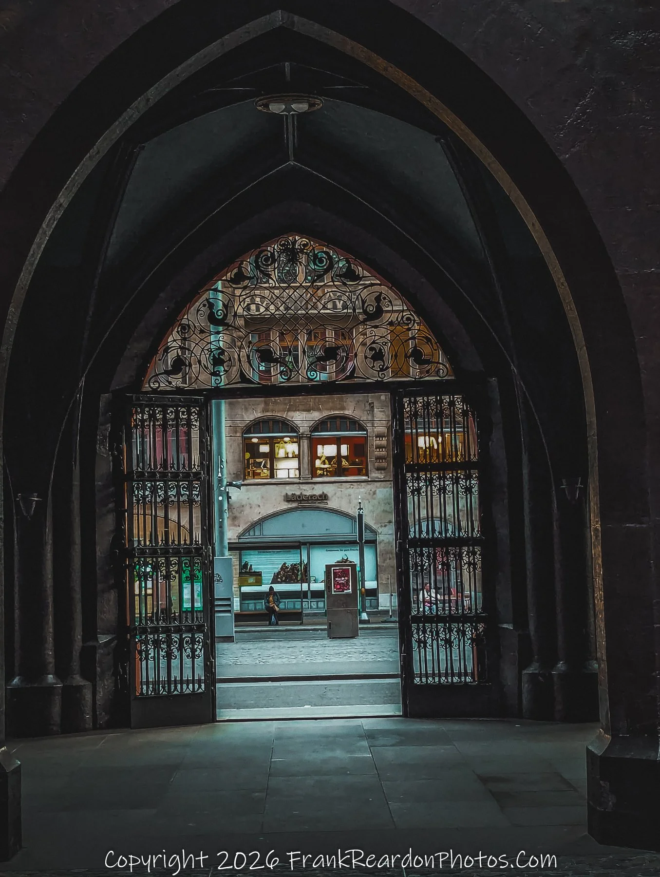 "Through the Iron Gates" A view looking out from the dark, arched stone entryway of the Rathaus toward the street beyond, framed by elaborate wrought-iron gates.