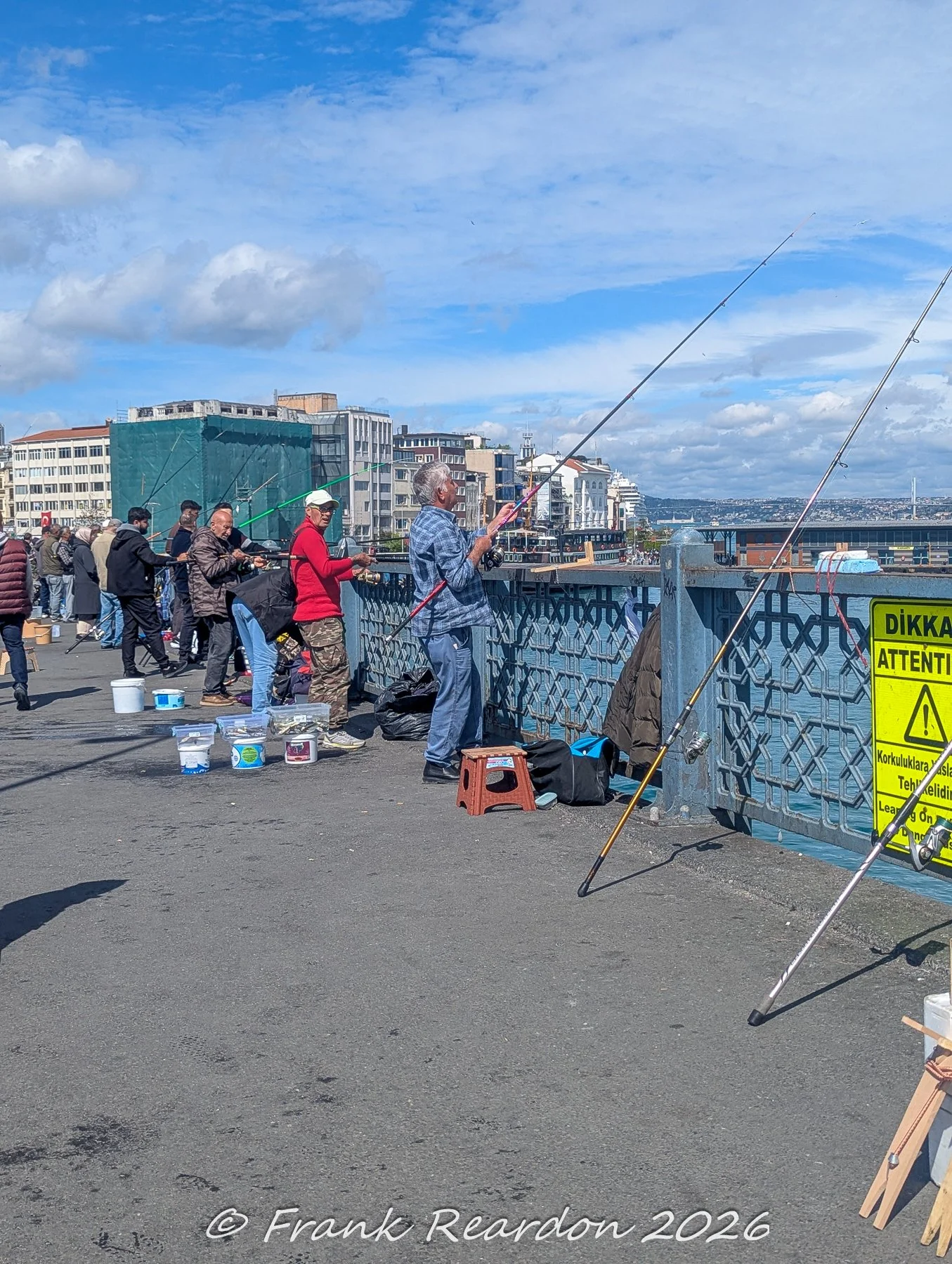 Local anglers lined up along the Galata Bridge with their fishing rods cast over the water, alongside a warning sign in Turkish and English.