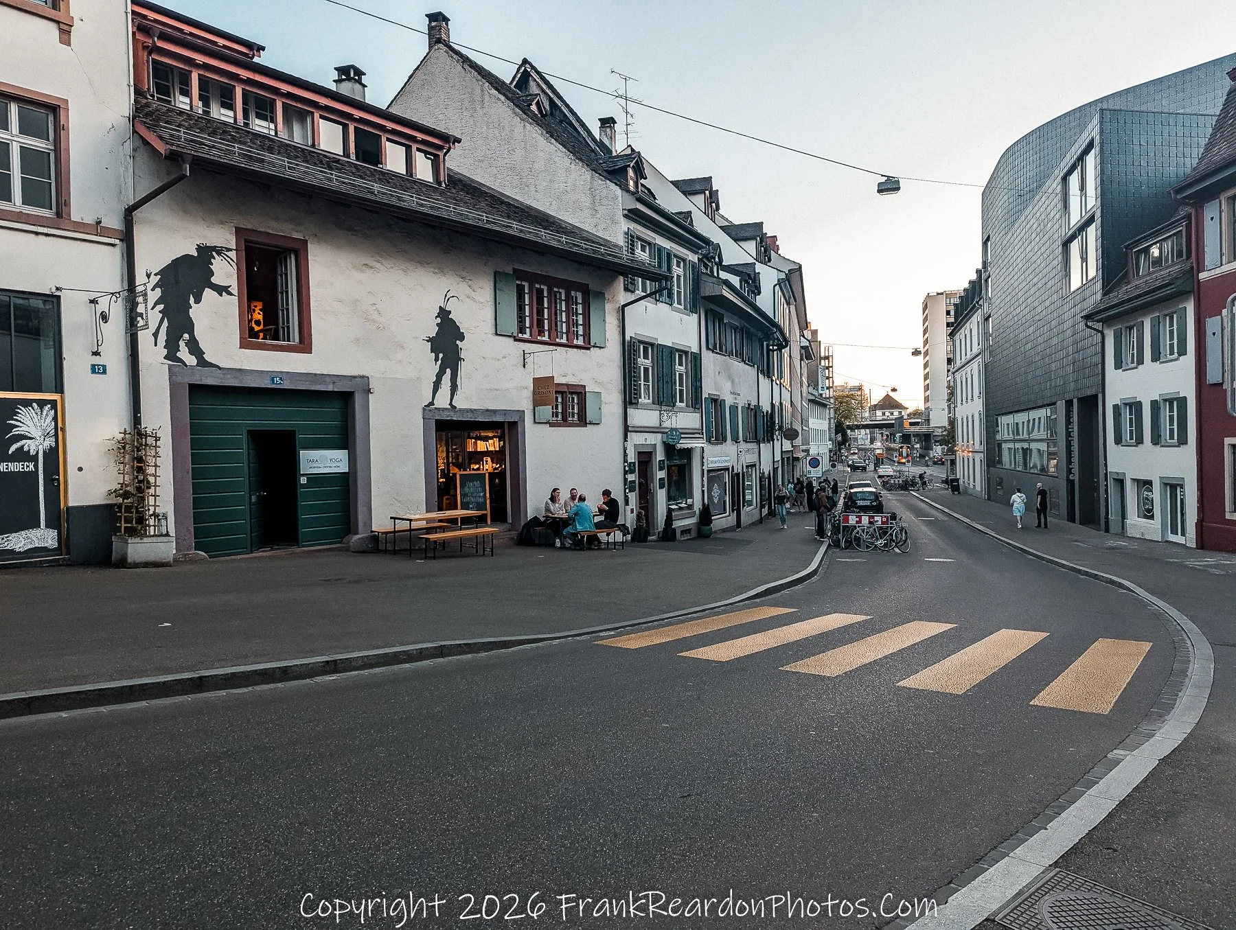 "Street View with Silhouettes" A quiet city street corner featuring white buildings adorned with black silhouette figures on the walls and outdoor dining tables.