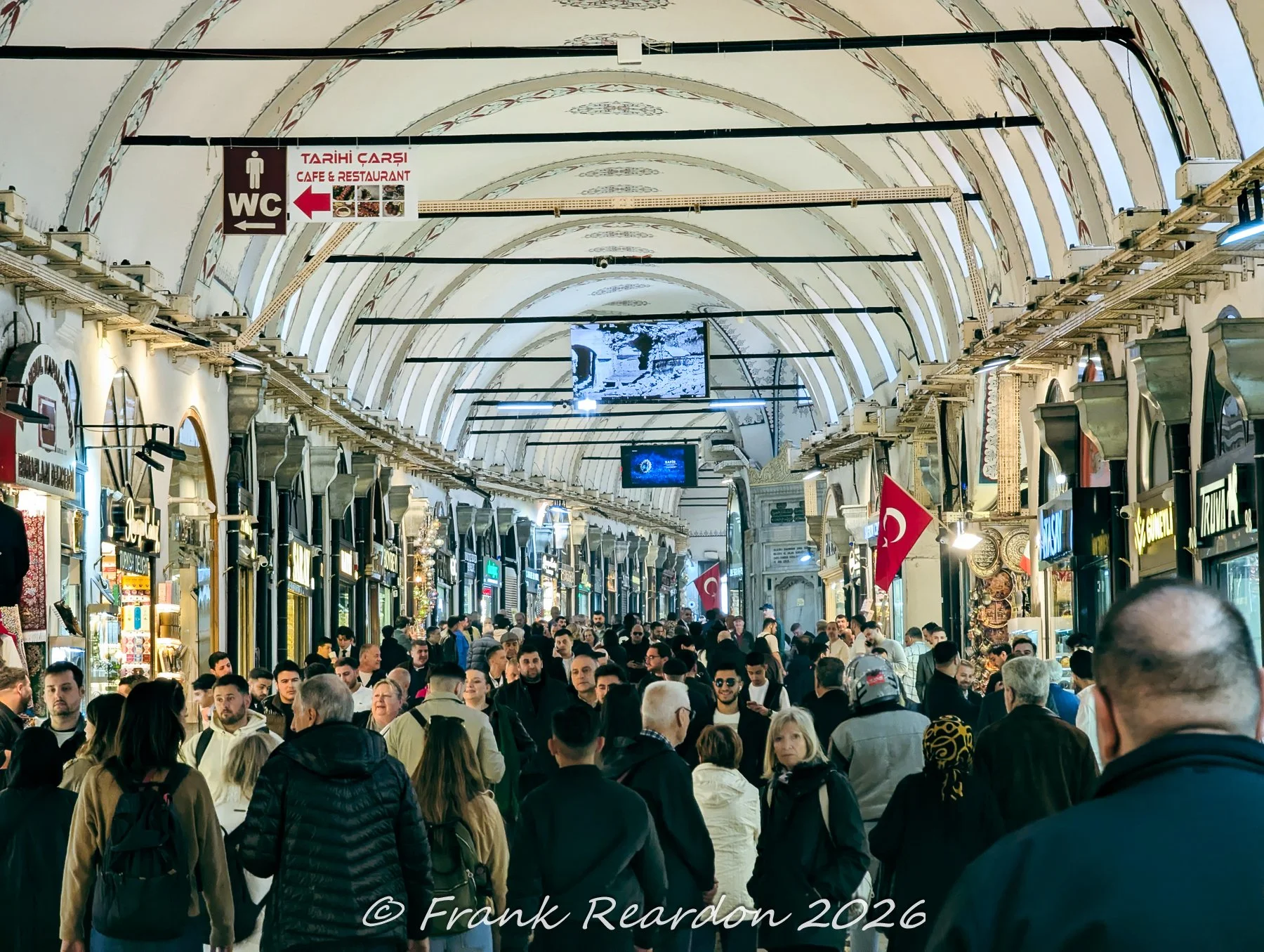 A lively scene filled with shoppers walking through the covered, vaulted arcade of the Grand Bazaar.