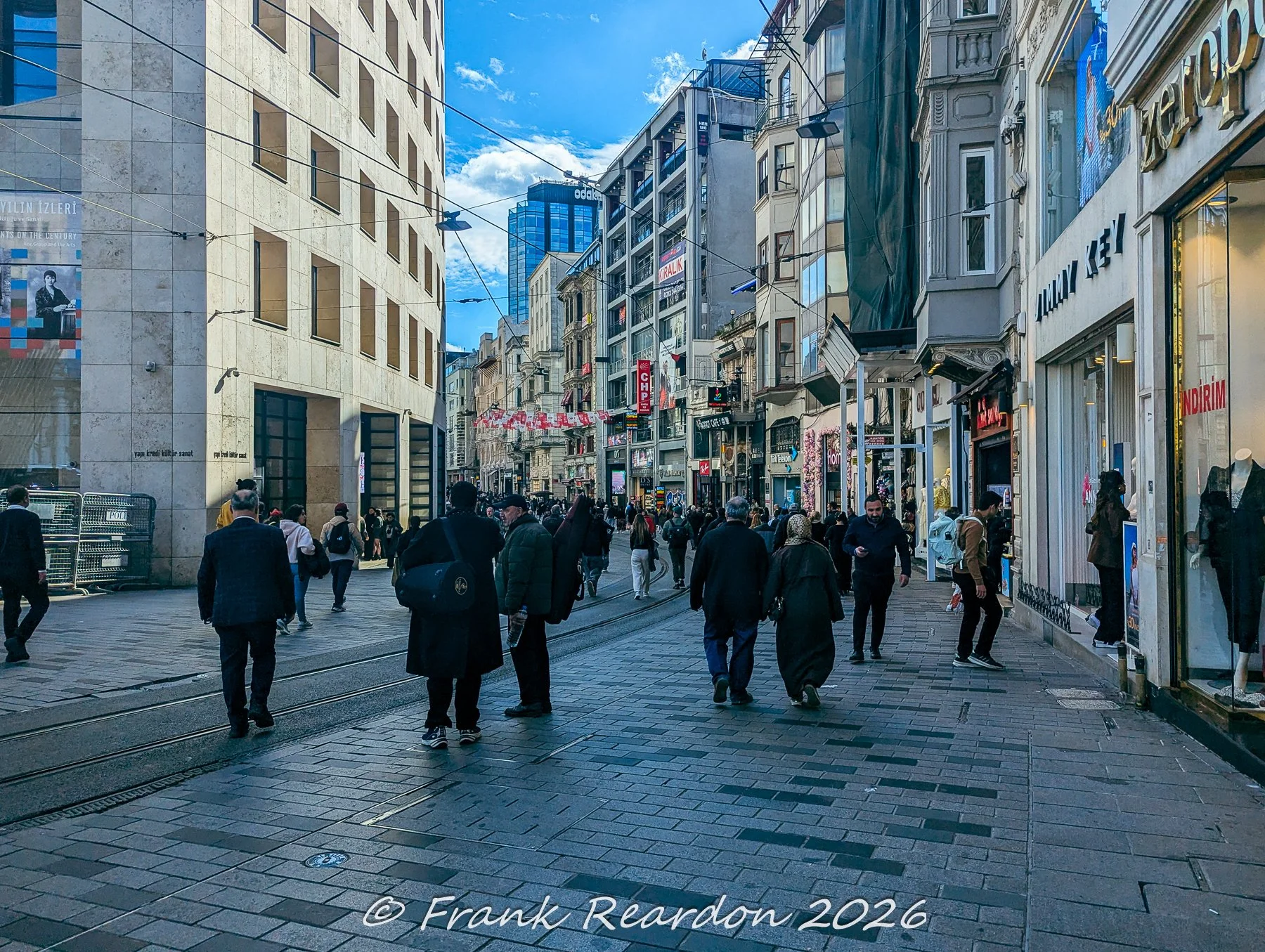 People walking along the lively, shop-lined corridor of Istiklal Avenue beneath an open blue sky and overhead power lines.