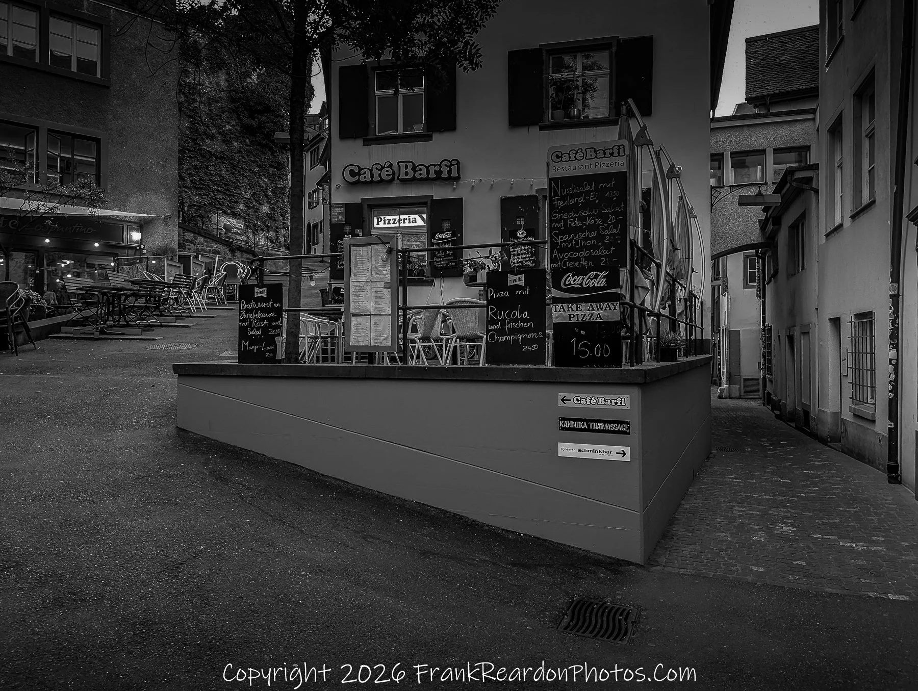 "Café Barfi Corner" A black-and-white view of Café Barfi, featuring outdoor menu boards listing various salads and pizzas, positioned at a street intersection.
