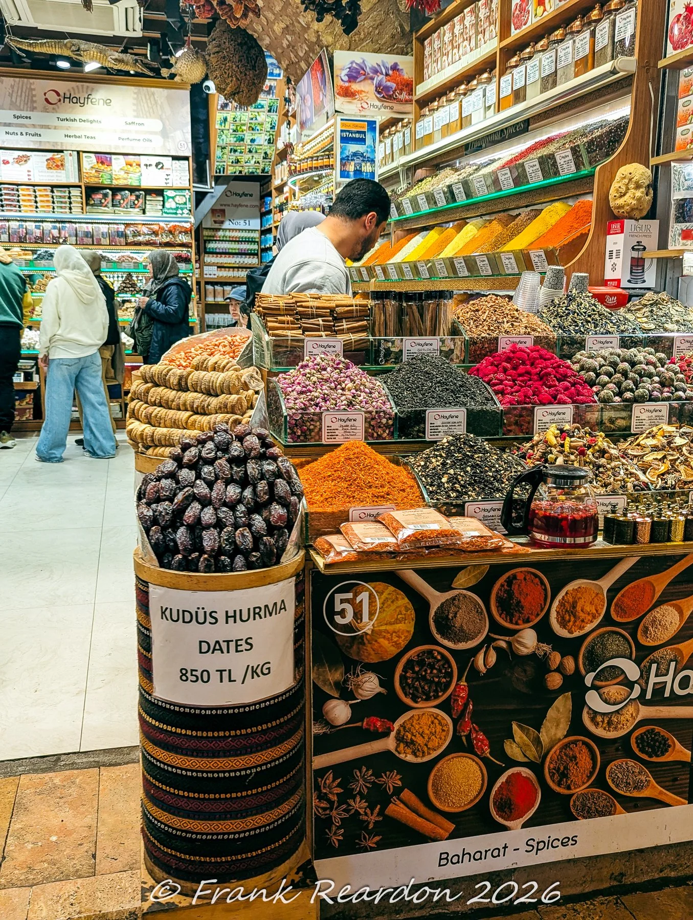 Market stalls showcasing large barrels of Kudüs Hurma dates, colorful dried botanicals, and traditional spice assortments within the Spice Bazaar.