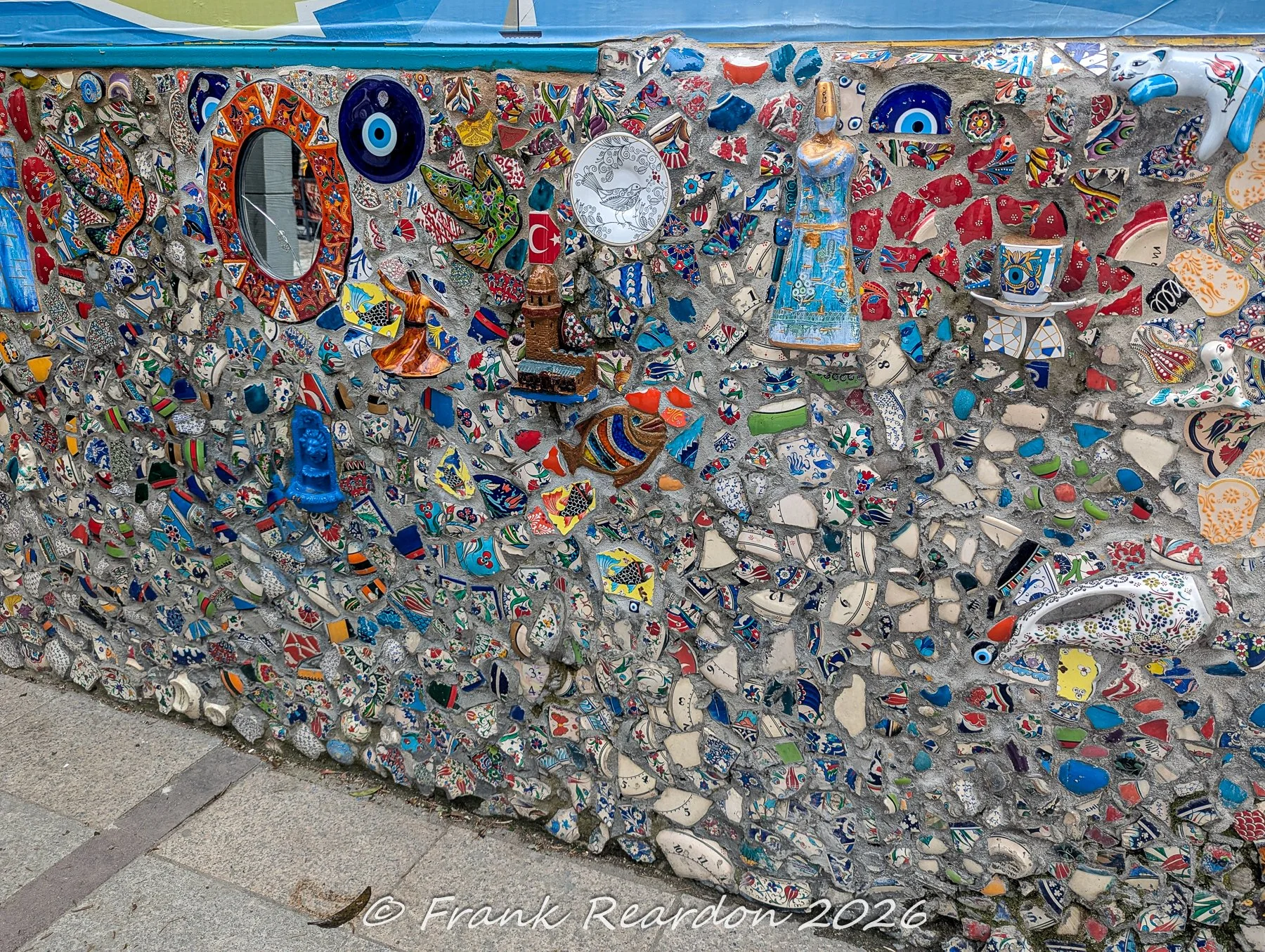 A detailed shot of an outdoor mosaic art installation embedded into a stone wall featuring broken ceramics, Turkish tiles, blue evil eye amulets, and traditional motifs.