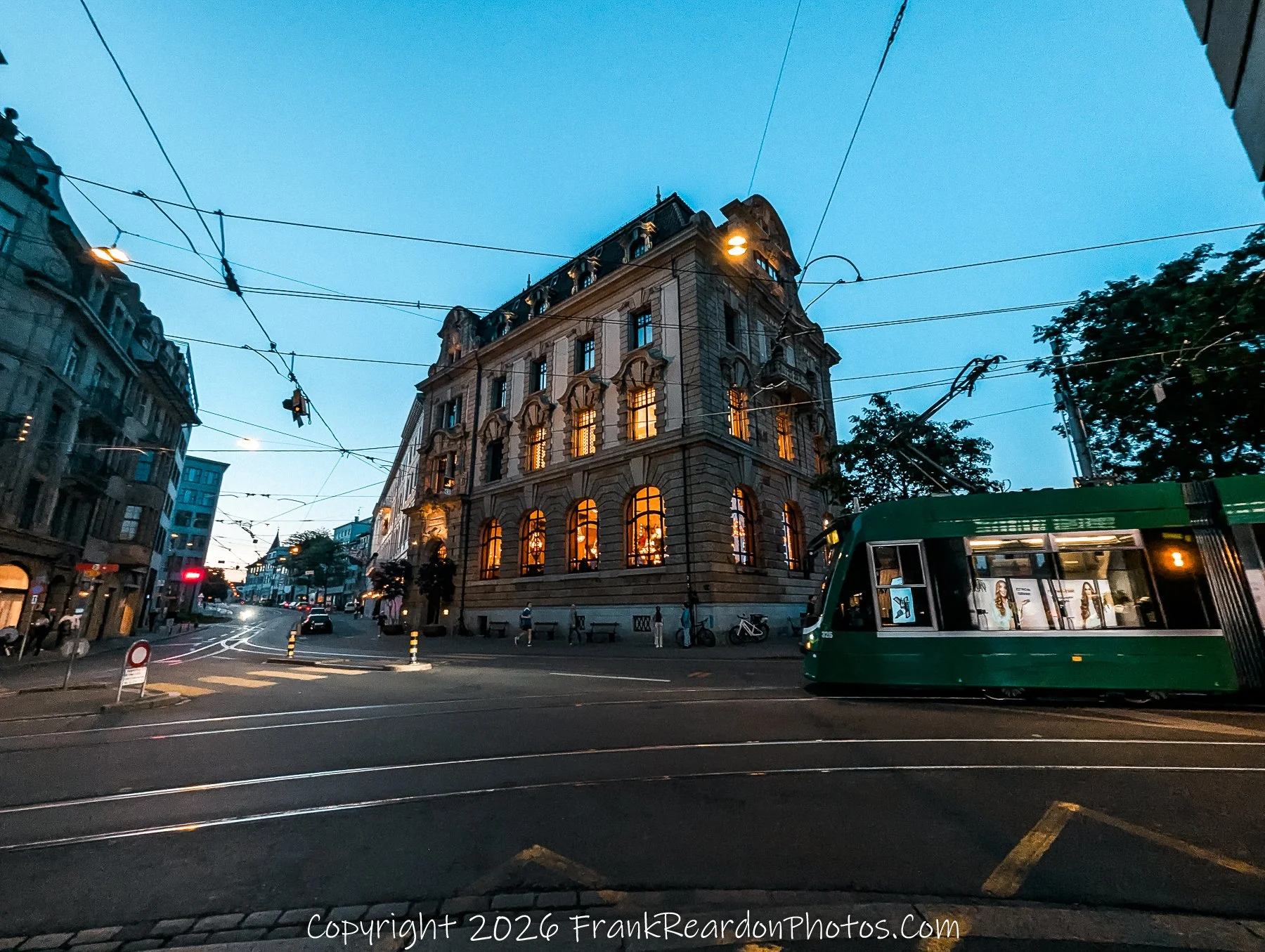 "Nighttime Commute" A green tram passing through a quiet intersection at dusk, set against the backdrop of an illuminated historic building.