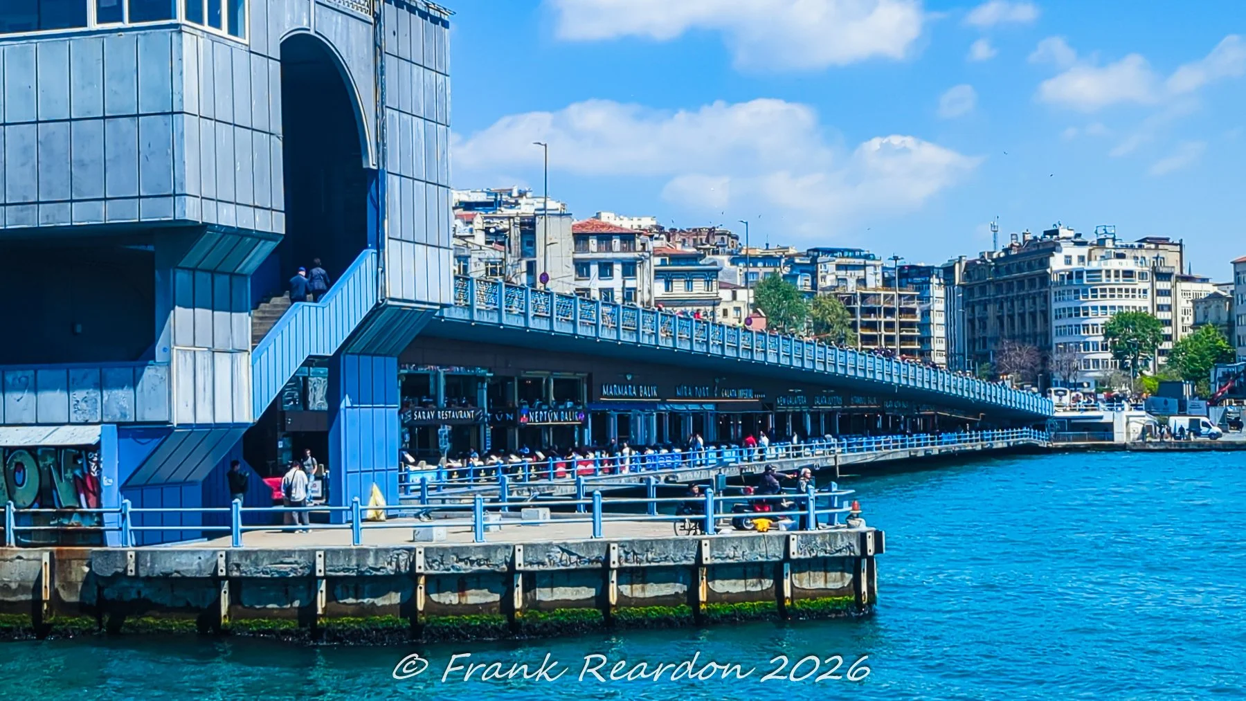 A vibrant shot showing the lower-level restaurants, walkways, and bustling waterfront cafes beneath the busy deck of the Galata Bridge spanning the Golden Horn.