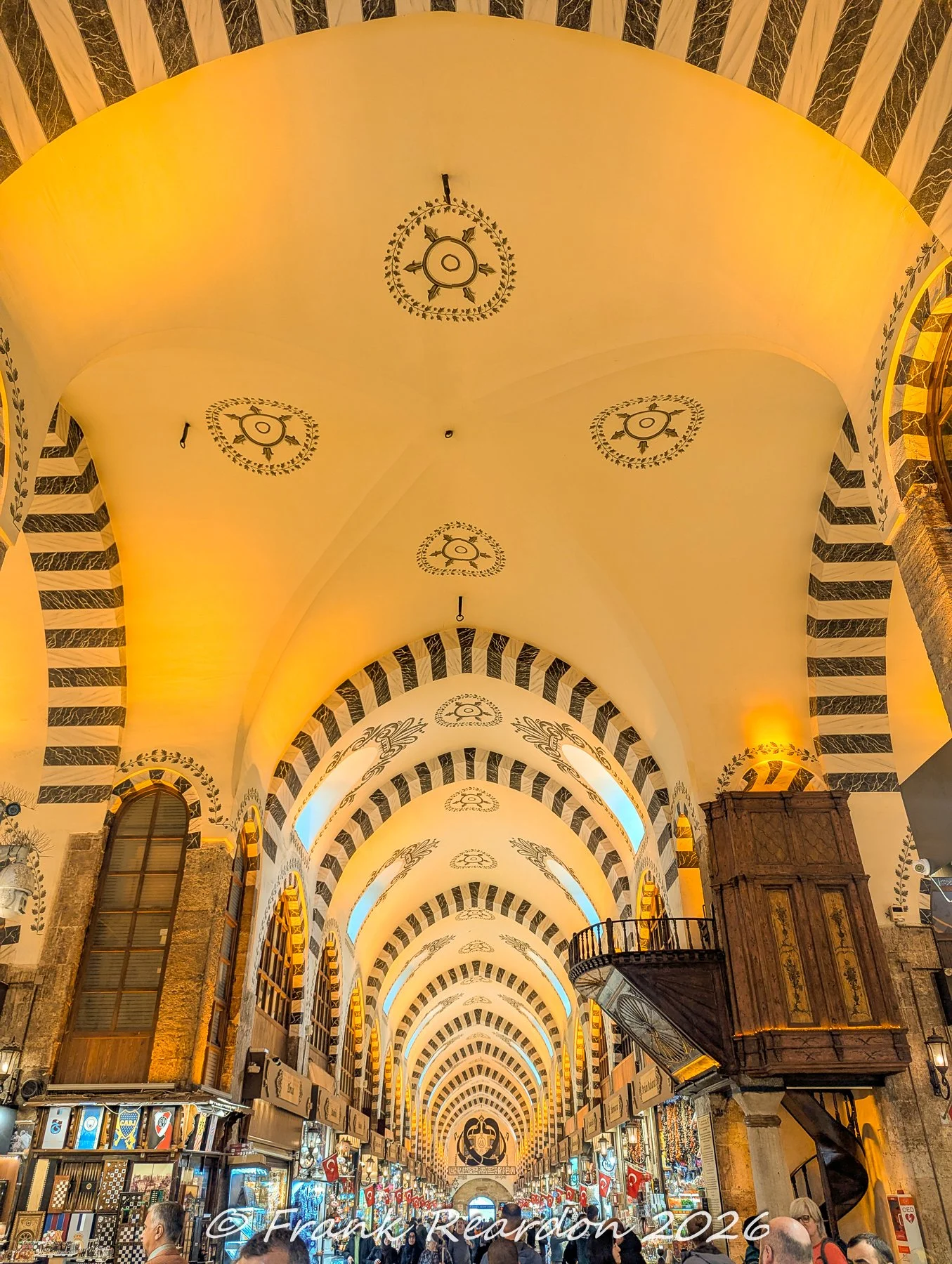 An impressive perspective looking down a long, whitewashed arcade featuring striped stone arches, decorative ceiling medallions, and a wooden pulpit structure inside the Grand Bazaar.