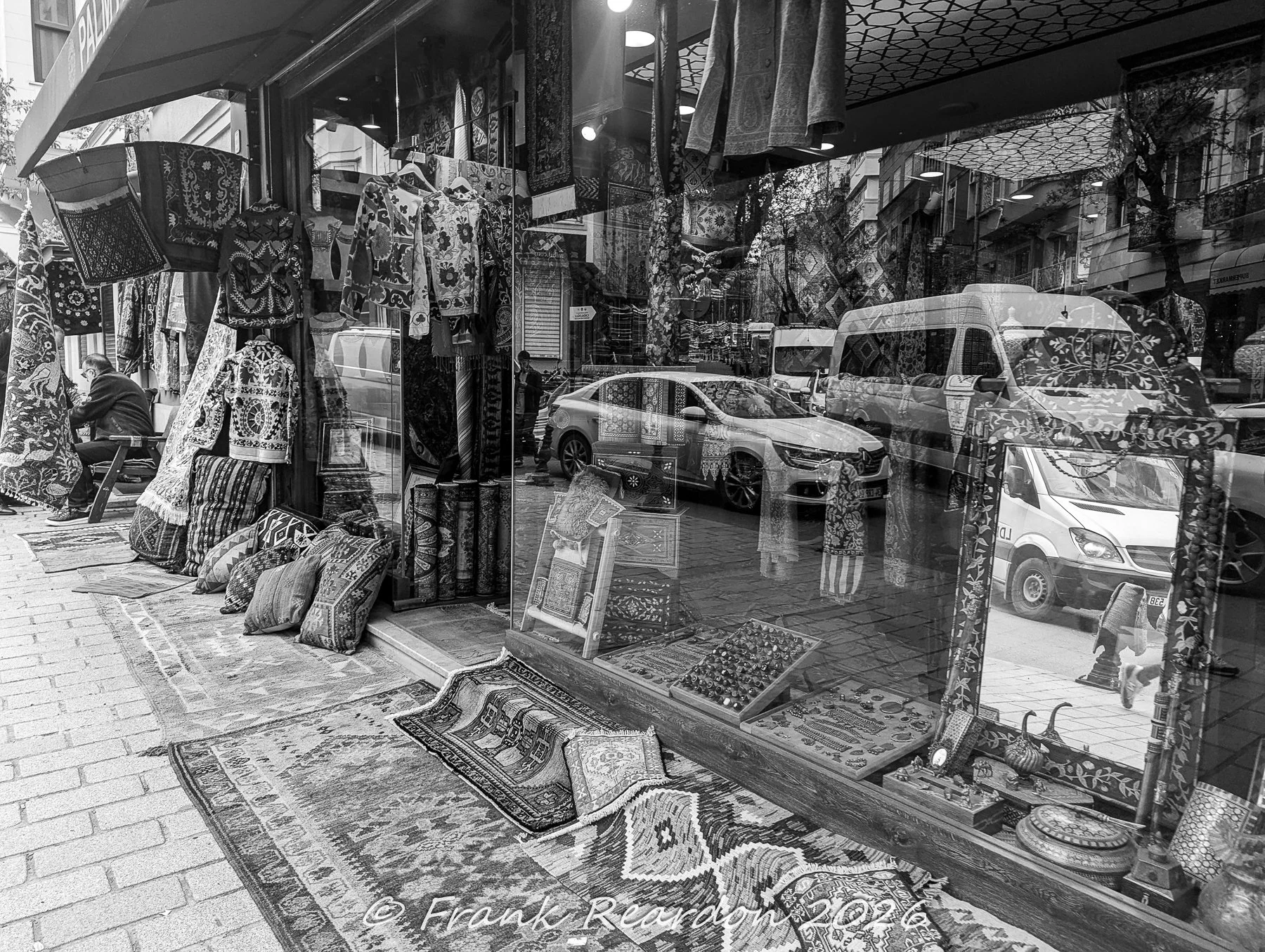 A black-and-white street scene reflecting passing cars and city life in the storefront window of an artisan shop displaying handmade rugs, pillows, and woven garments.
