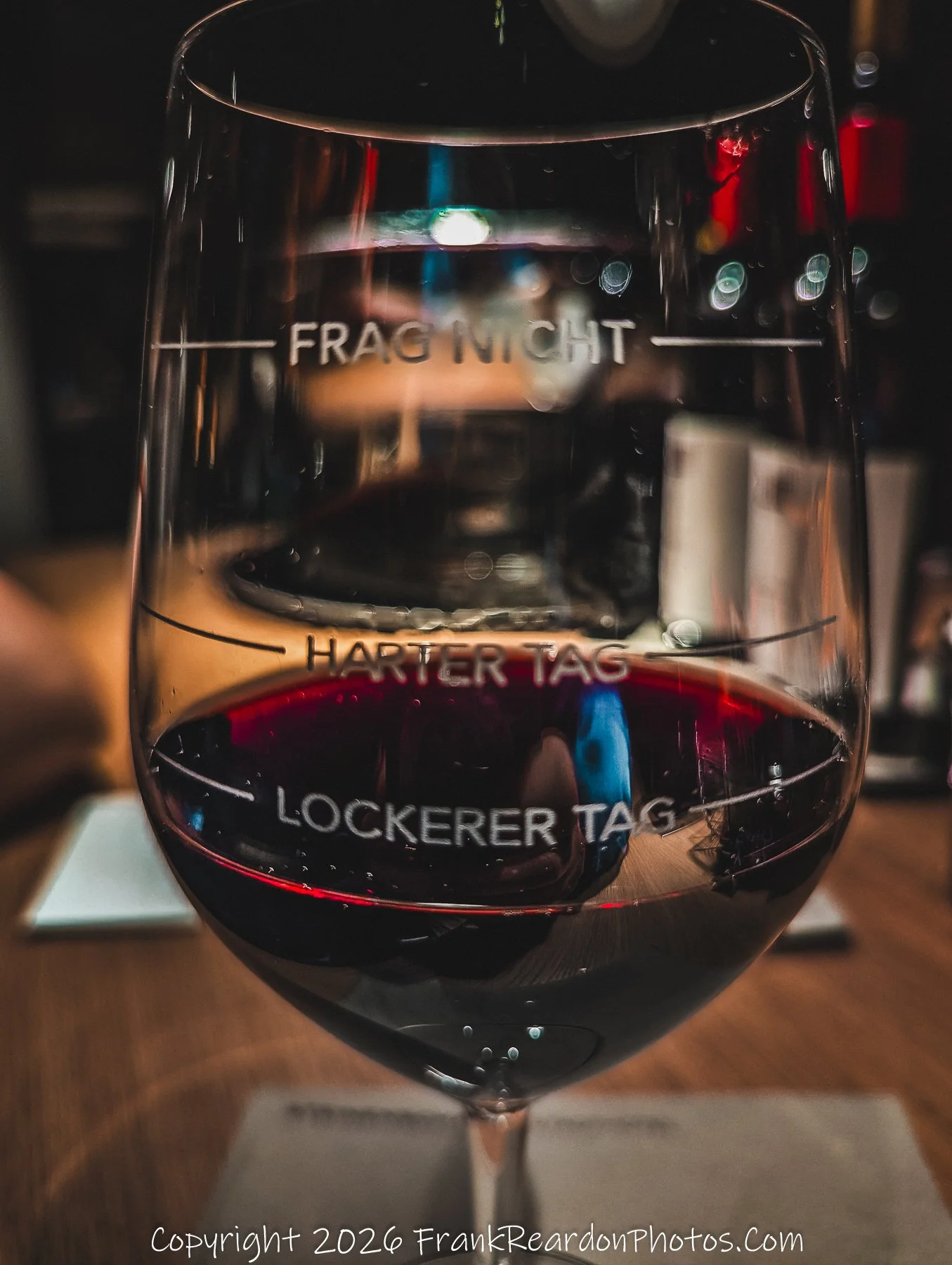 "A Measure of the Day" A creative close-up shot of a wine glass etched with humorous labels "Frag Nicht" (Don't Ask), "Harter Tag" (Hard Day), and "Lockerer Tag" (Easy Day) filled with red wine.
