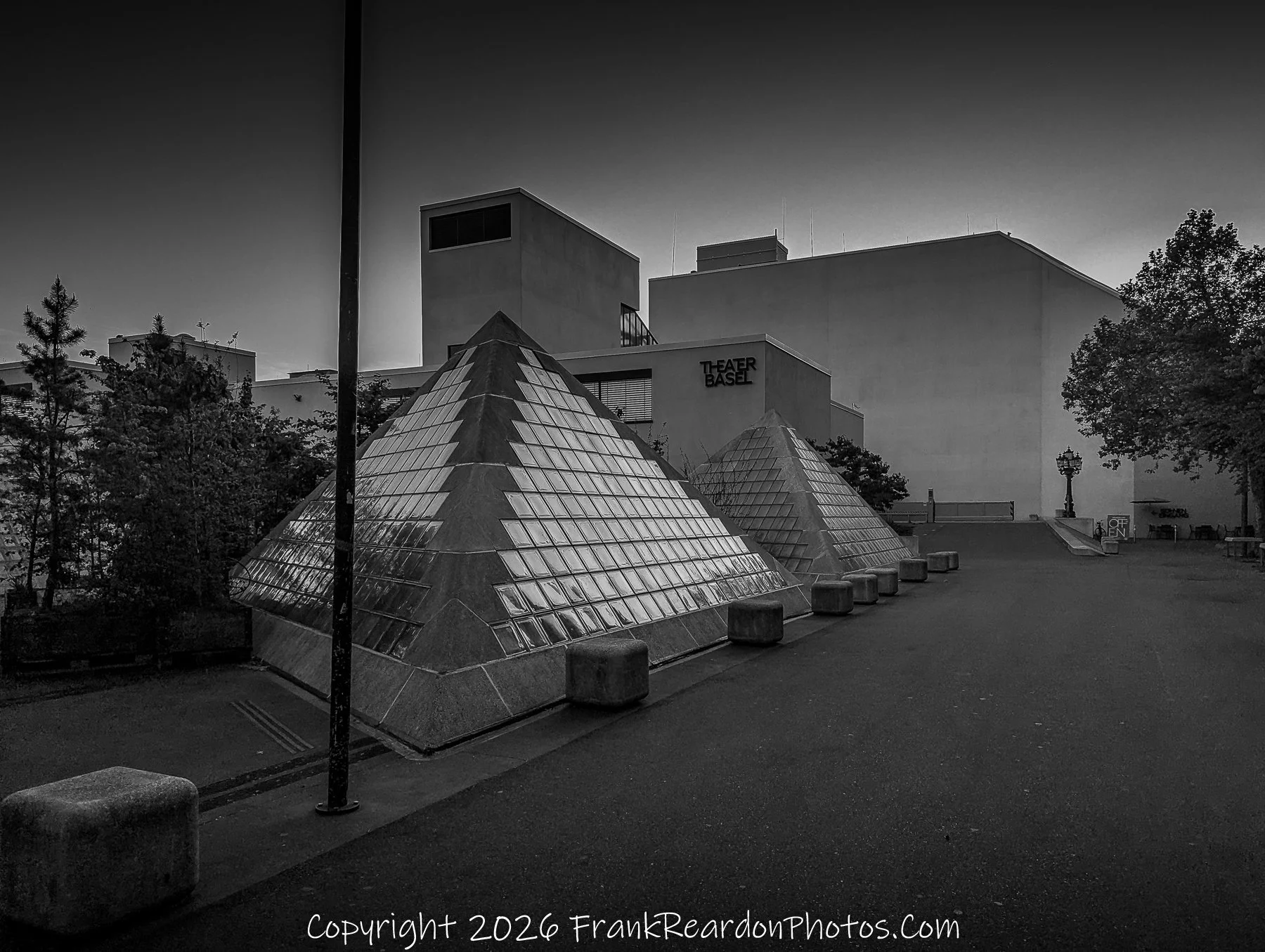 "Glass Pyramids at Twilight" A black-and-white image featuring two glass-paneled pyramids in a paved square, with the "Theater Basel" building visible in the background.