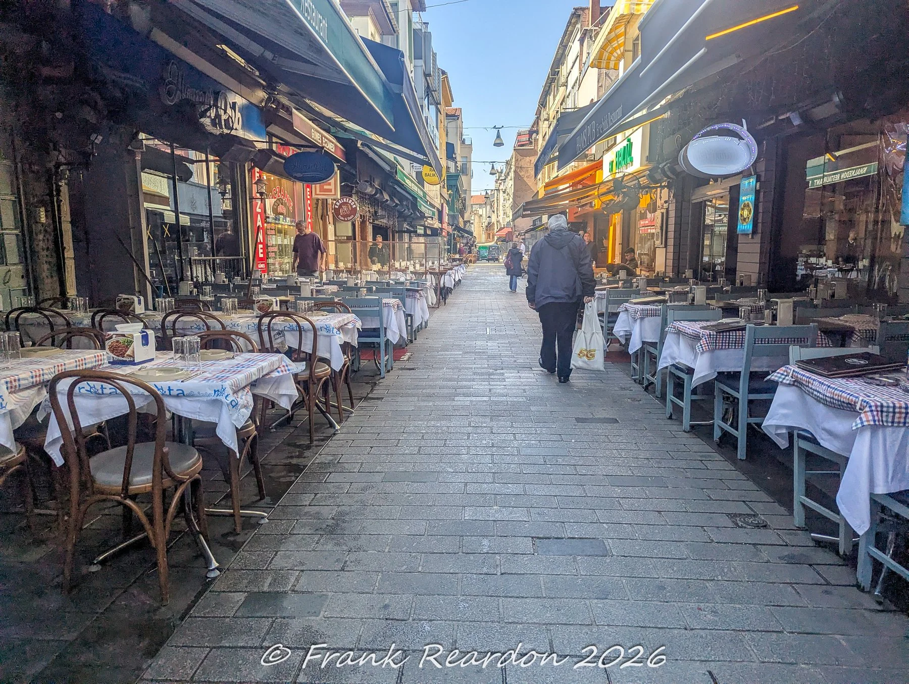 A daytime view down a narrow street lined with outdoor restaurant seating, checkered tablecloths, awnings, and a lone pedestrian walking with a bag.