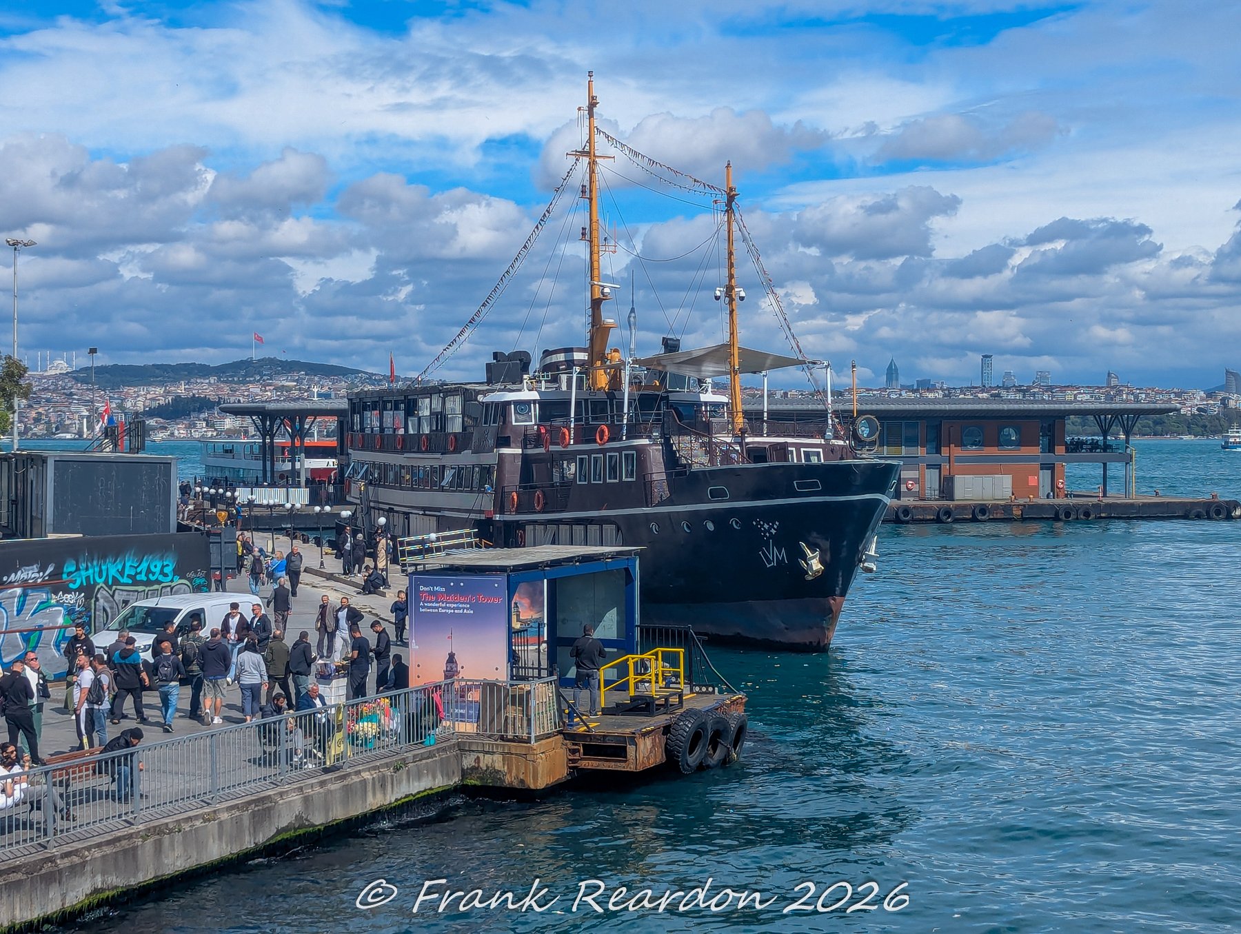 A historic dark-hulled passenger ferry docked at a modern terminal along the bustling Istanbul waterfront.