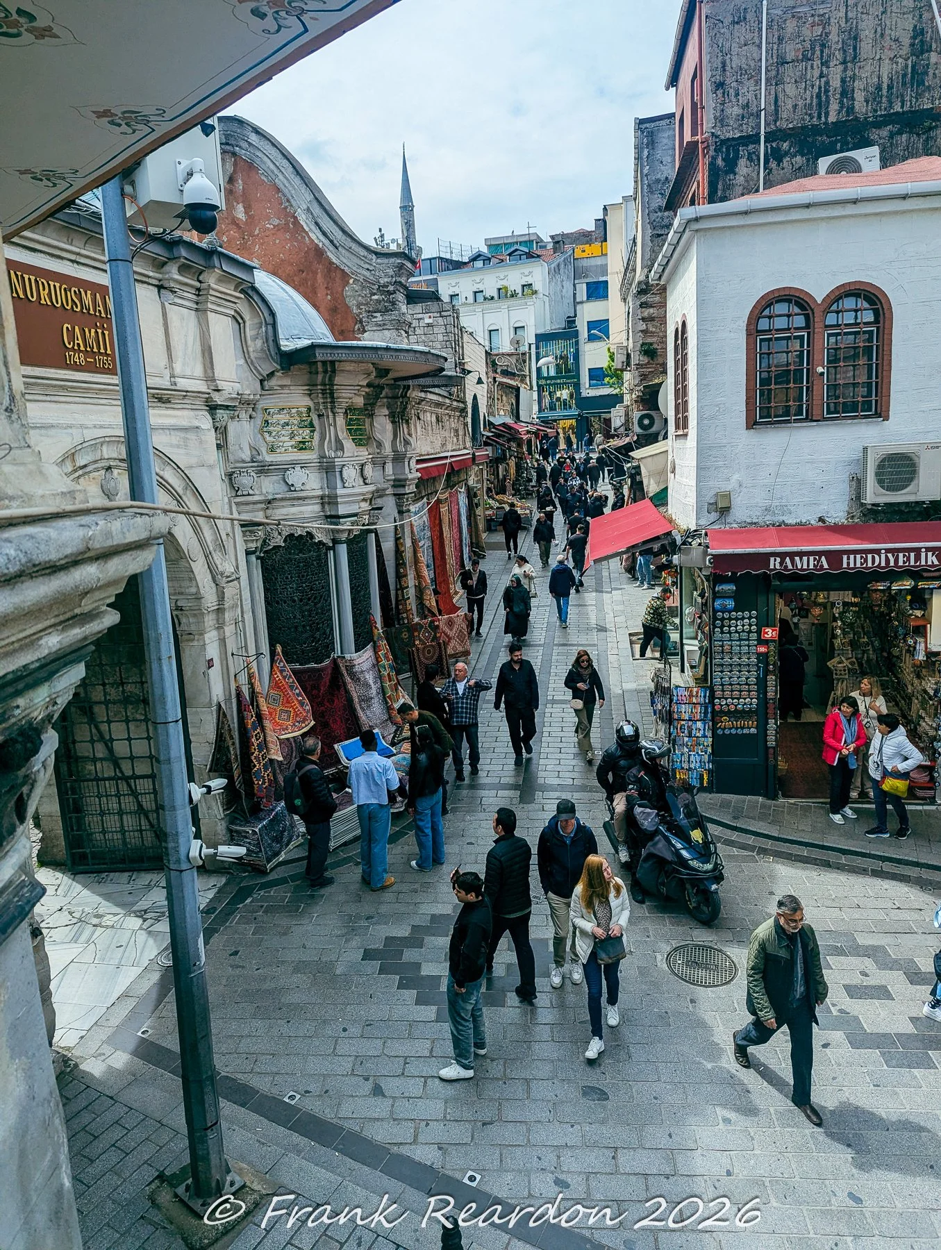 An elevated perspective looking down a lively historic street lined with shops, carpet displays, and pedestrians near the Nuruosmaniye Mosque.