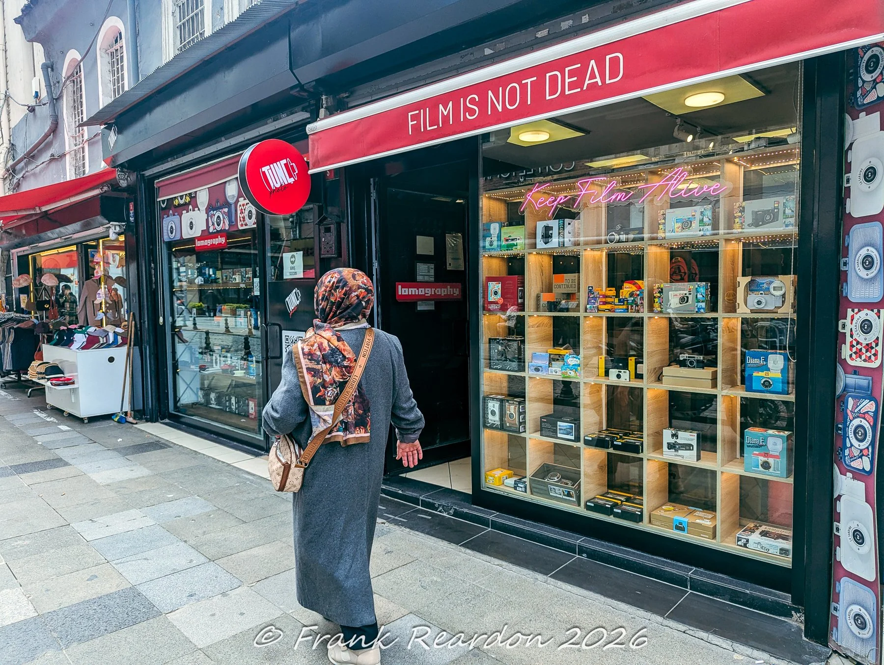 A woman walking past a specialized photography shop window filled with vintage cameras, film rolls, and Lomography gear on a city street.
