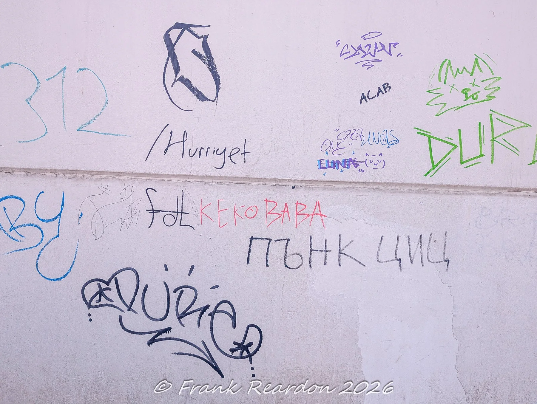 A detailed look at a white-painted wall covered in various overlapping street tags, marker signatures, and multilingual graffiti.