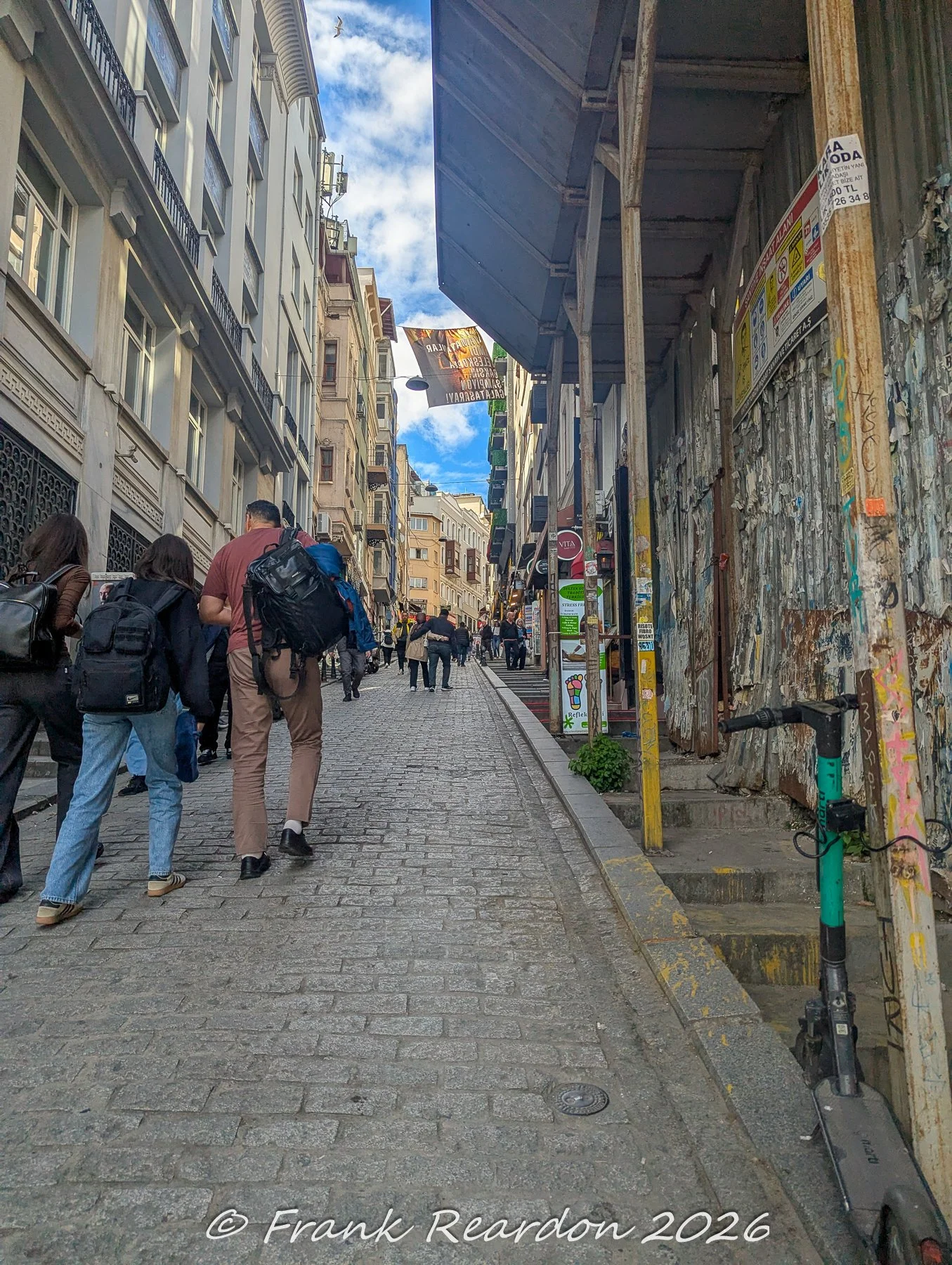 Pedestrians walking up a steep, narrow cobblestone street lined with multi-story European-style buildings, scaffolding, and a parked scooter.