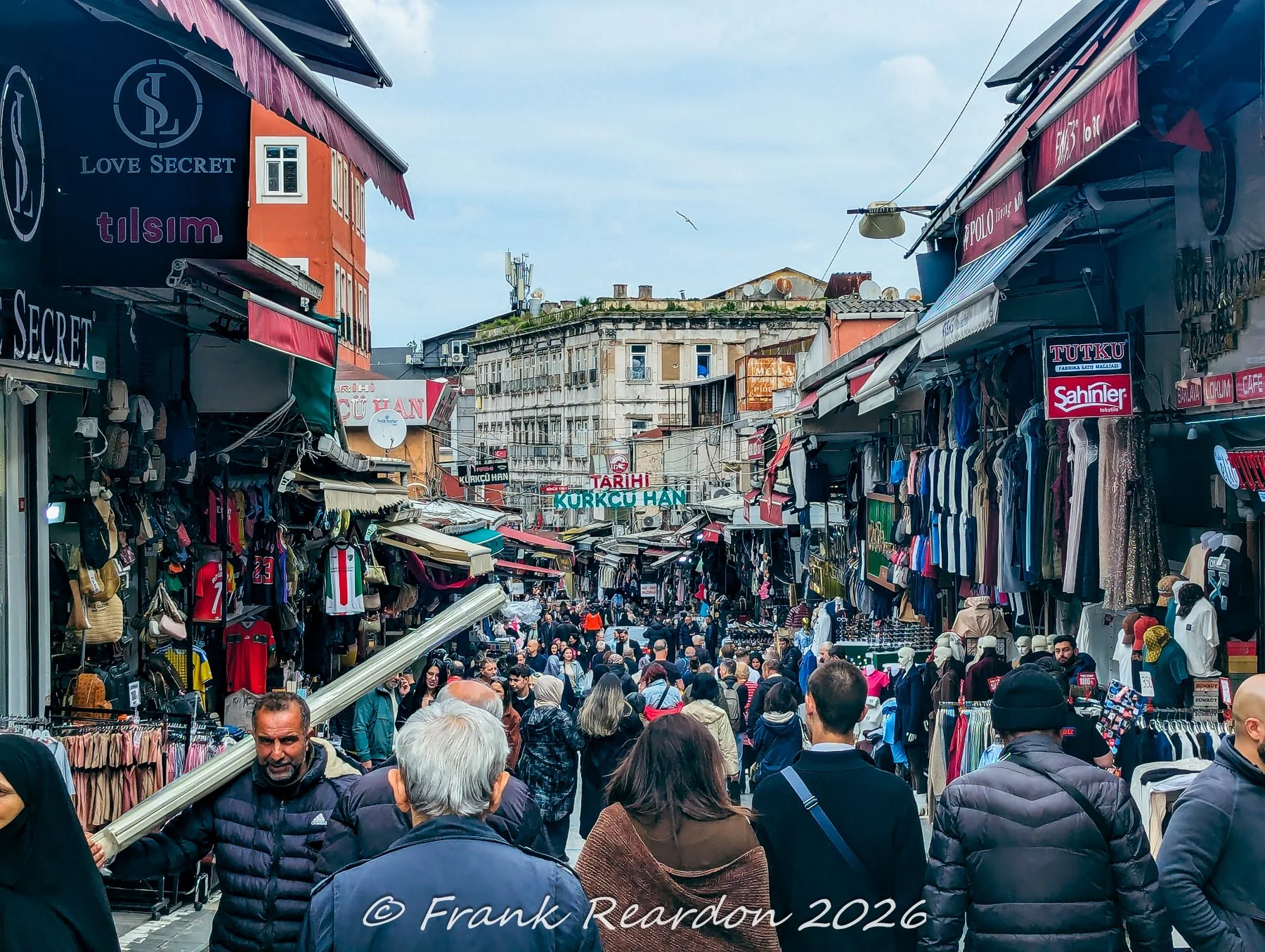 Shoppers and pedestrians strolling through a busy outdoor market alley lined with clothing vendors, awnings, and historic buildings near the Grand Bazaar.