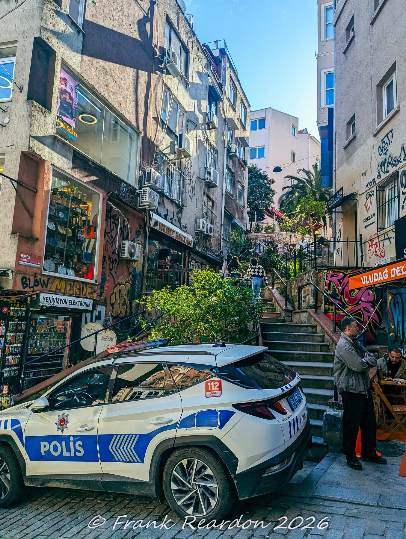 A sloping urban street featuring a parked Turkish police vehicle, stone staircases leading upward, and colorful graffiti near local shops.