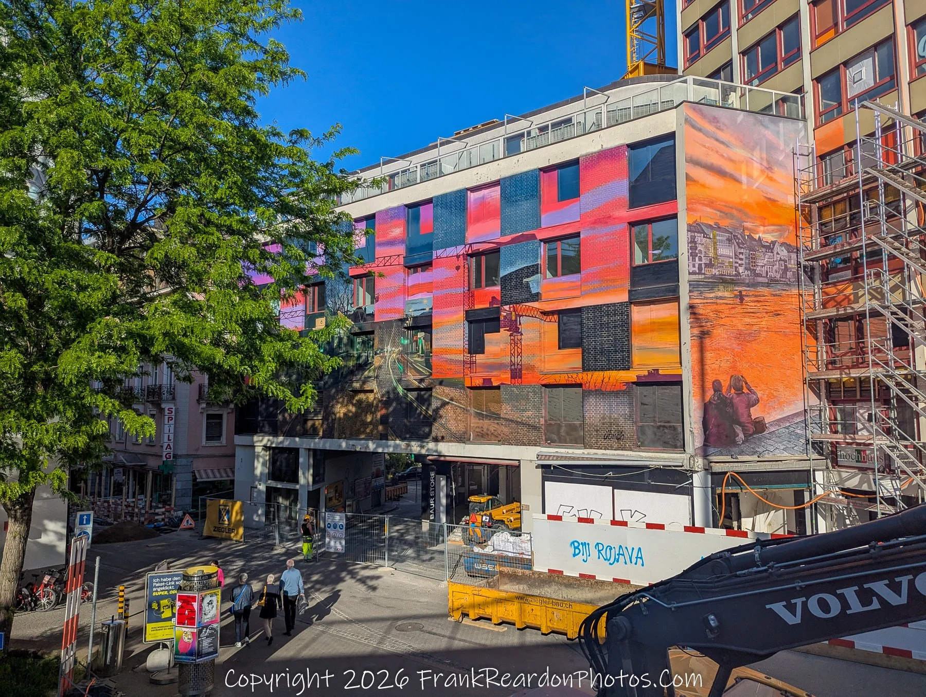 "Sunset Mural in the City" A large-scale mural painted on the side of a multi-story building, depicting a vivid sunset over a waterfront, located near an active construction site with a large crane and a dumpster labeled "Biji Rojava."