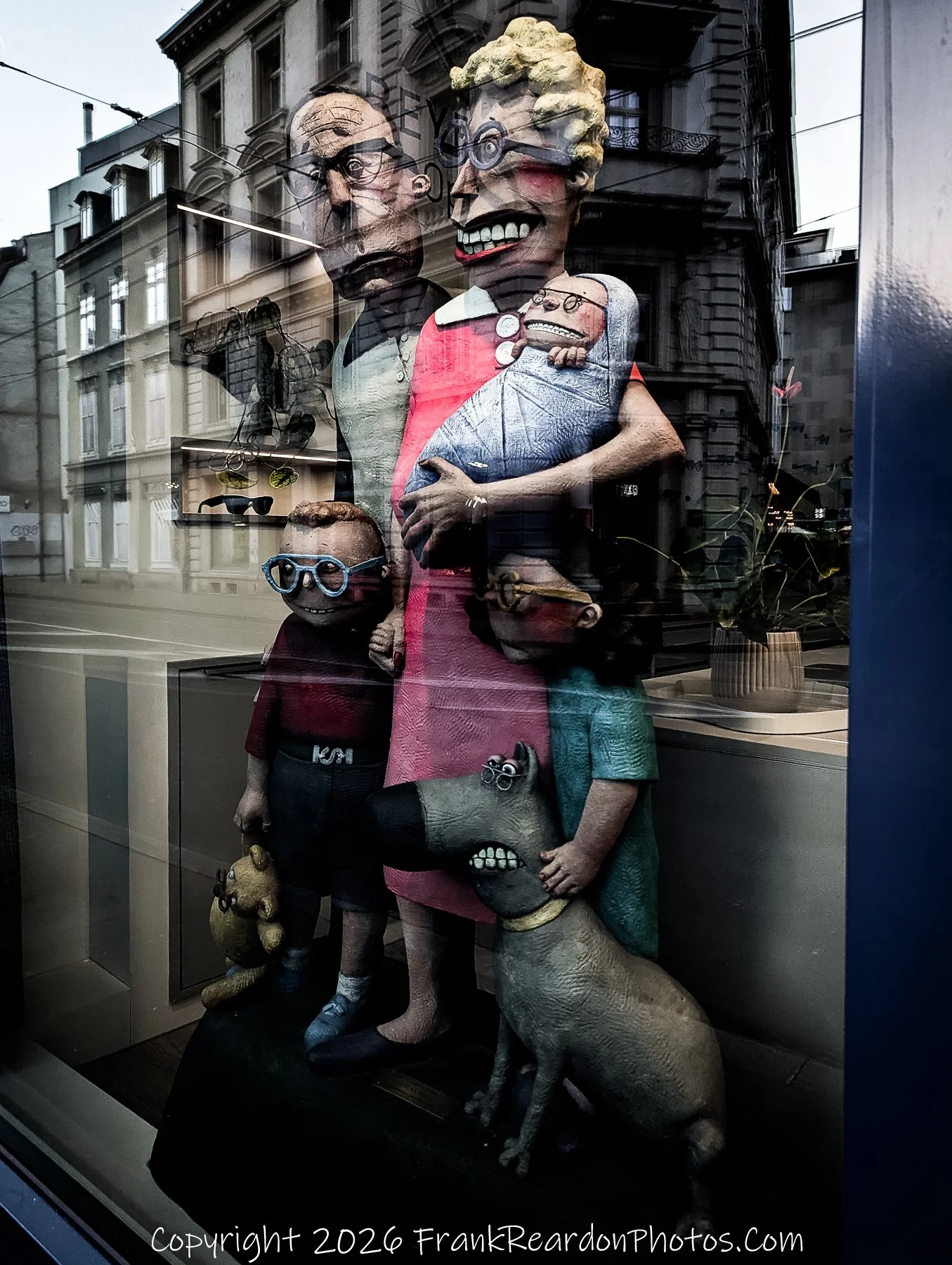 "Whimsical Family Statue" A store window display featuring stylized, caricatured figures of a family and a dog, with their reflections visible in the glass.