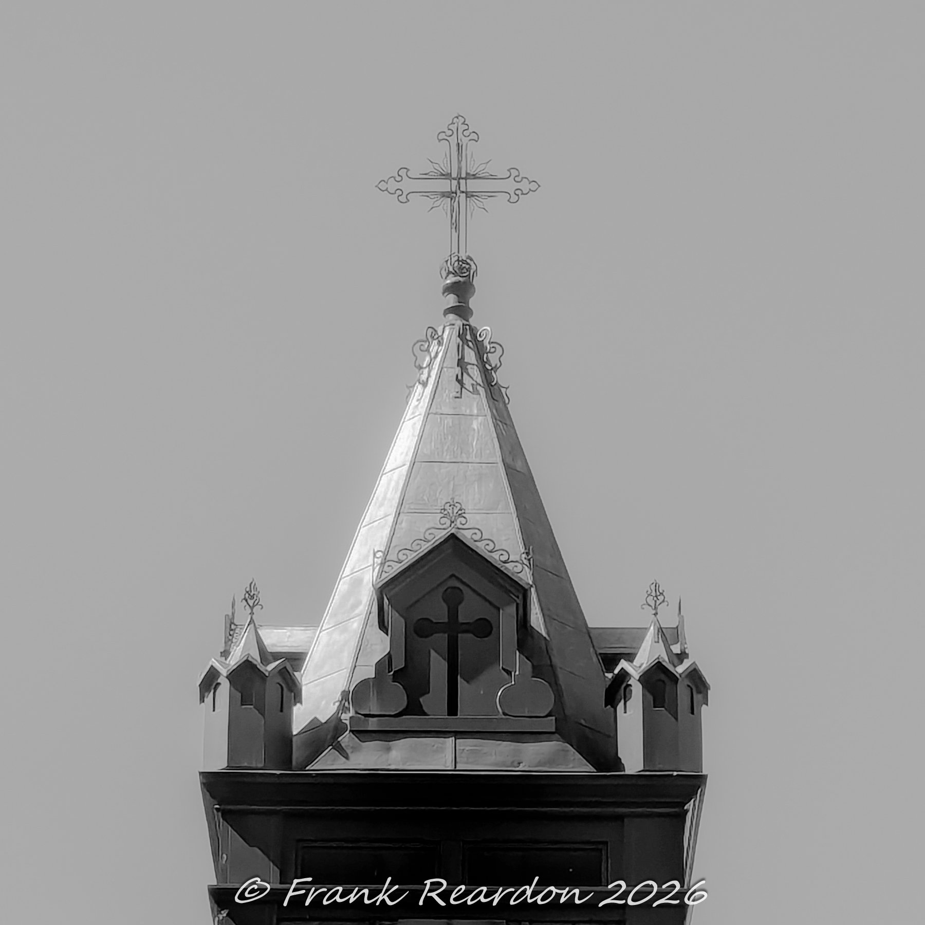 A detailed black-and-white architectural shot focusing on the pointed roof, small corner turrets, and intricate cross atop a church tower.