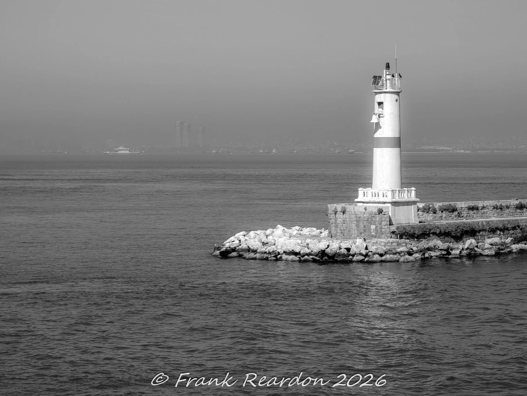 A black-and-white coastal view of a white lighthouse with a red stripe standing on a rocky outcrop at the edge of the Bosphorus.   