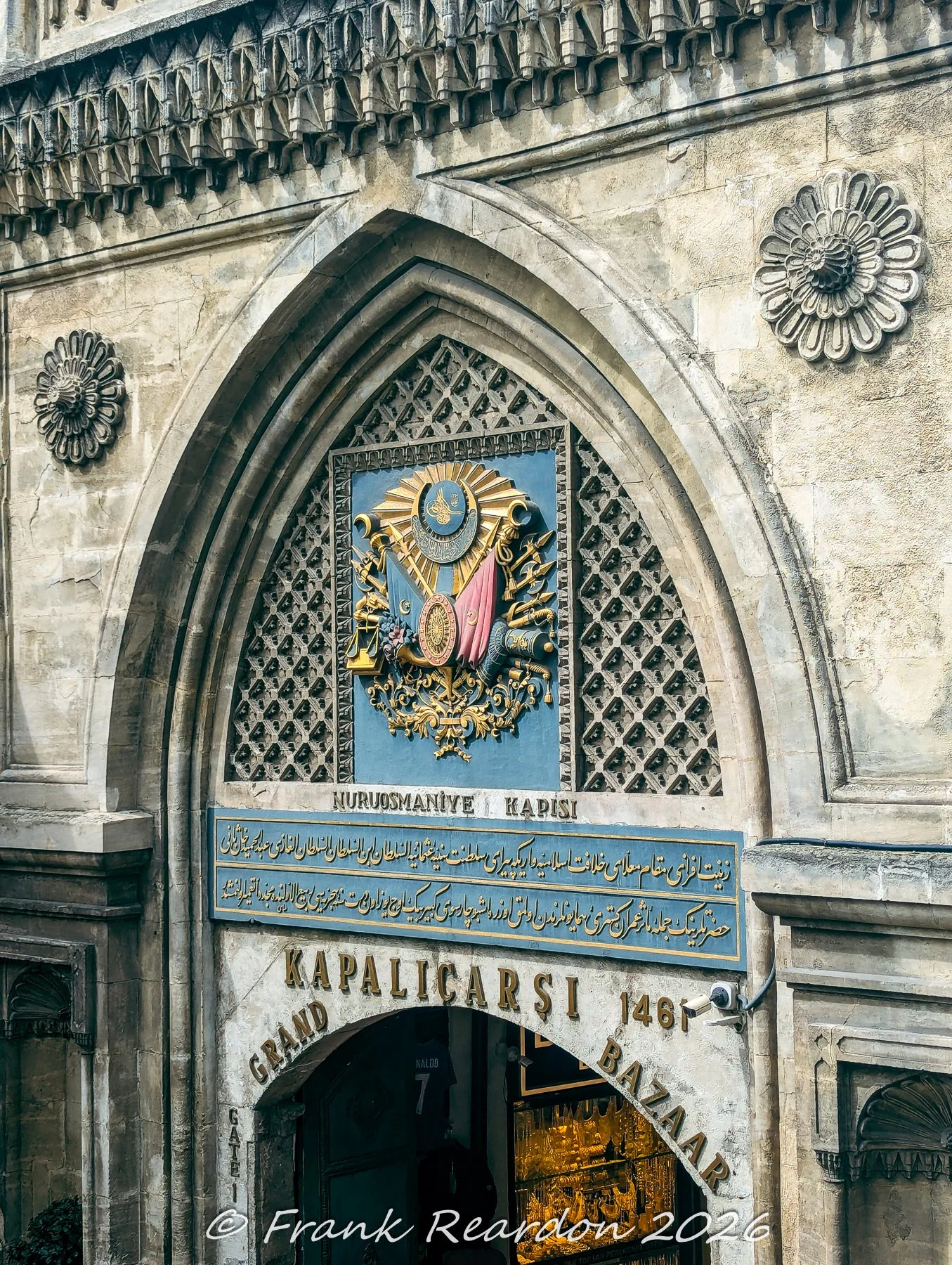 A close-up view of the majestic stone archway featuring the Ottoman coat of arms, the "Nuruosmaniye Kapısı" sign, and the entrance to the Grand Bazaar (Kapalıçarşı).