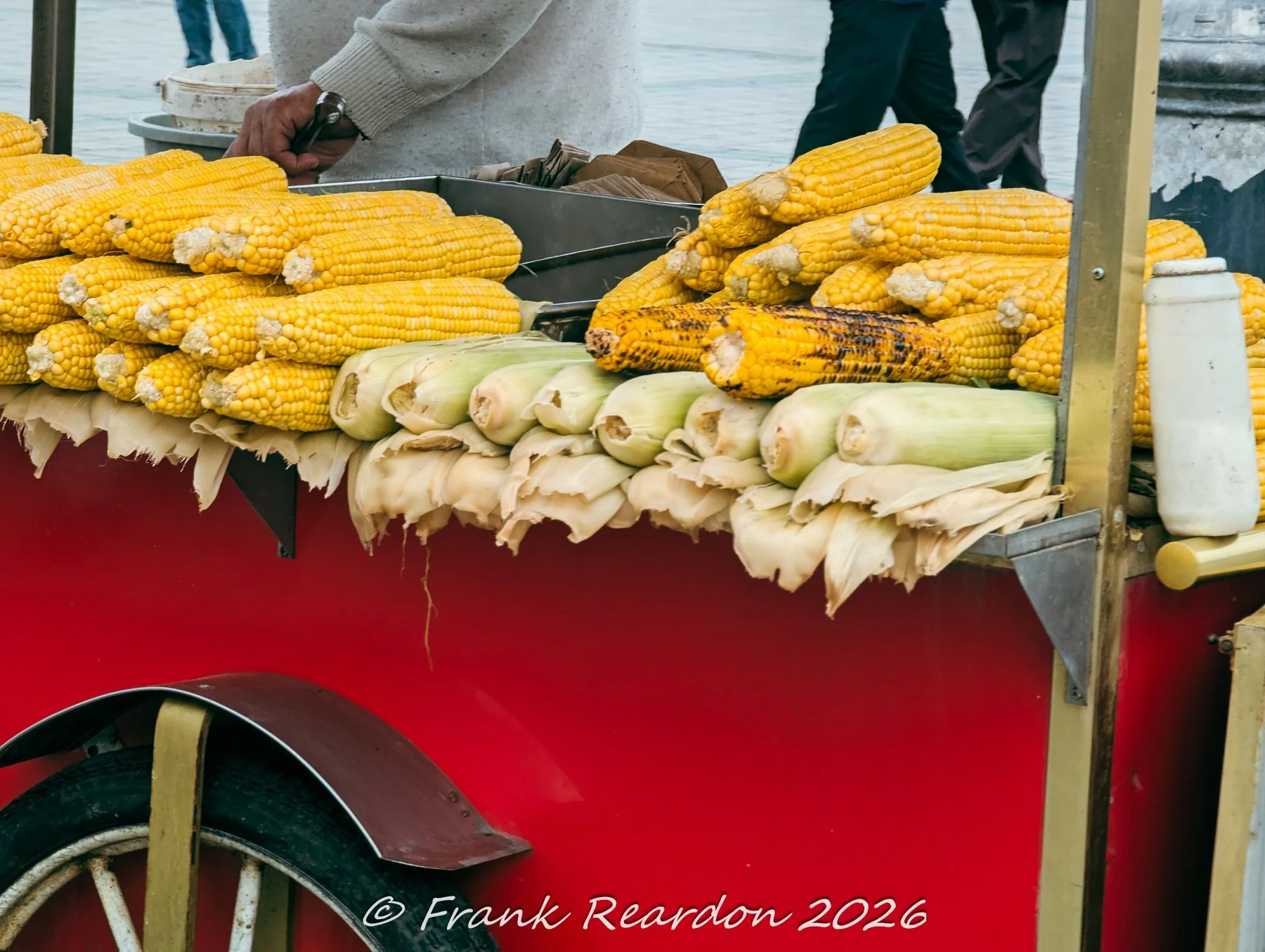 A traditional red street cart loaded with piles of golden boiled corn on the cob and husked raw corn cobs on a street in Istanbul.