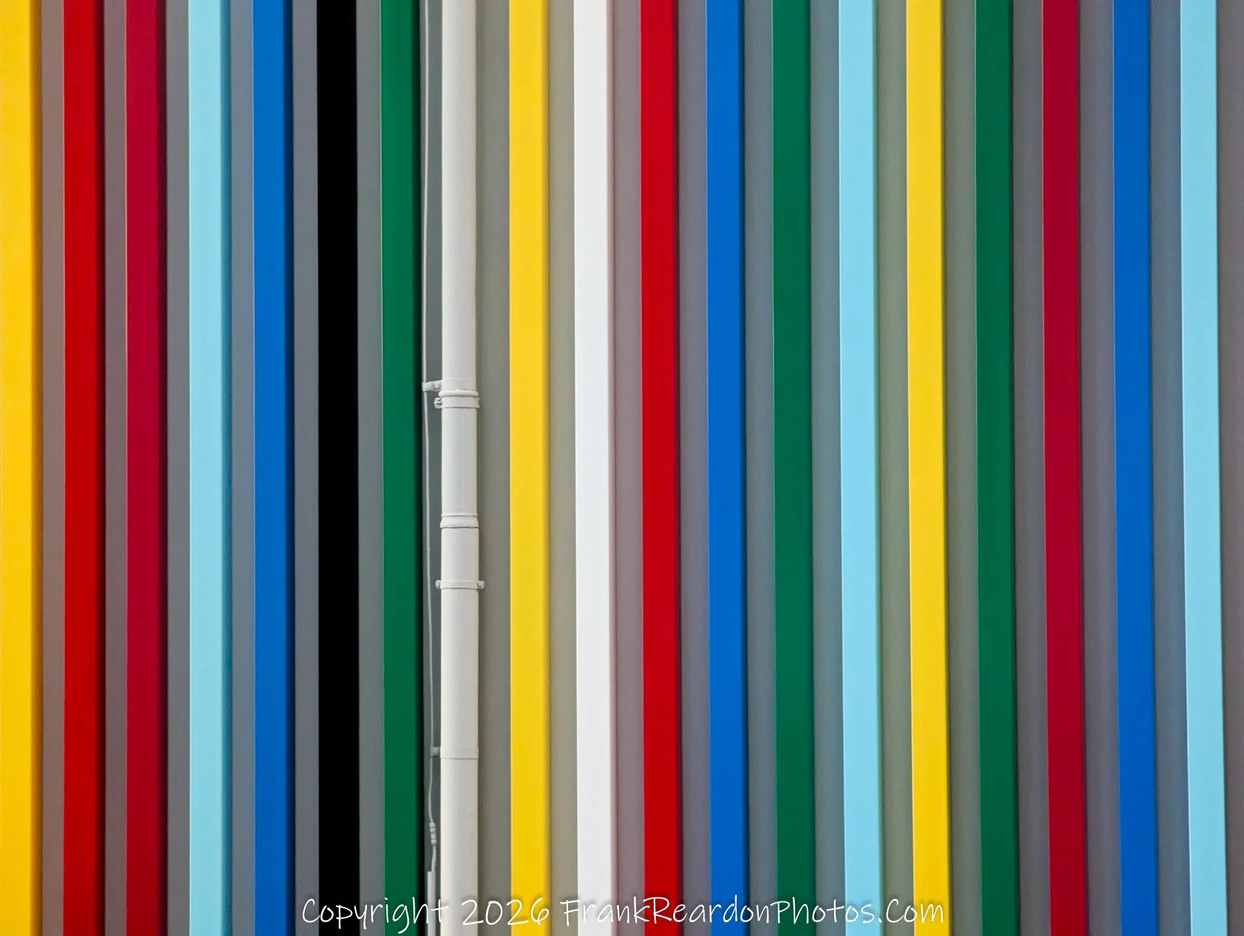 "Abstract Vertical Stripes" A close-up view of the same multi-colored architectural wall, showing a white vertical utility pipe running down the center of the striped pattern.