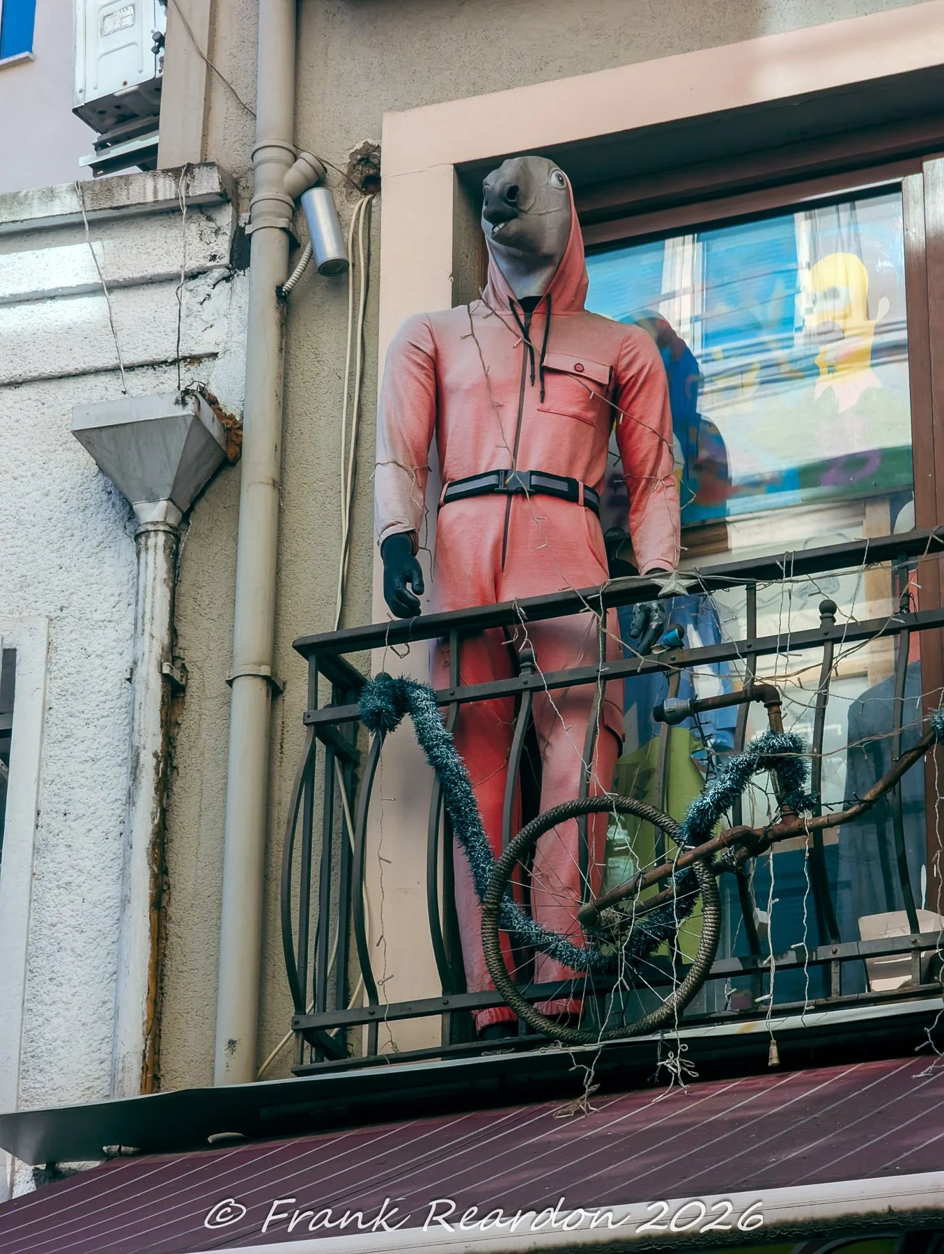 A mannequin wearing a pink jumpsuit with an animal mask, posed on an apartment balcony alongside a bicycle frame.